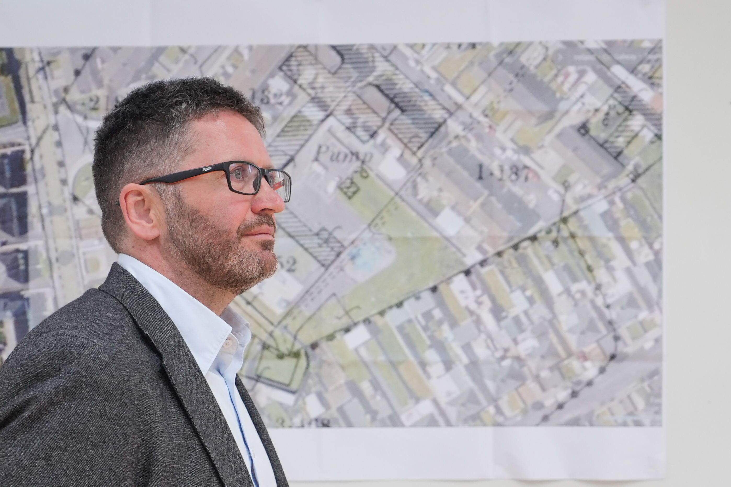 Un hombre con chaqueta gris, camisa clara y gafas mira hacia un lado. Tiene el pelo corto y oscuro con canas. Está de pie frente a un mapa topográfico superpuesto a la imagen de un yacimiento.
