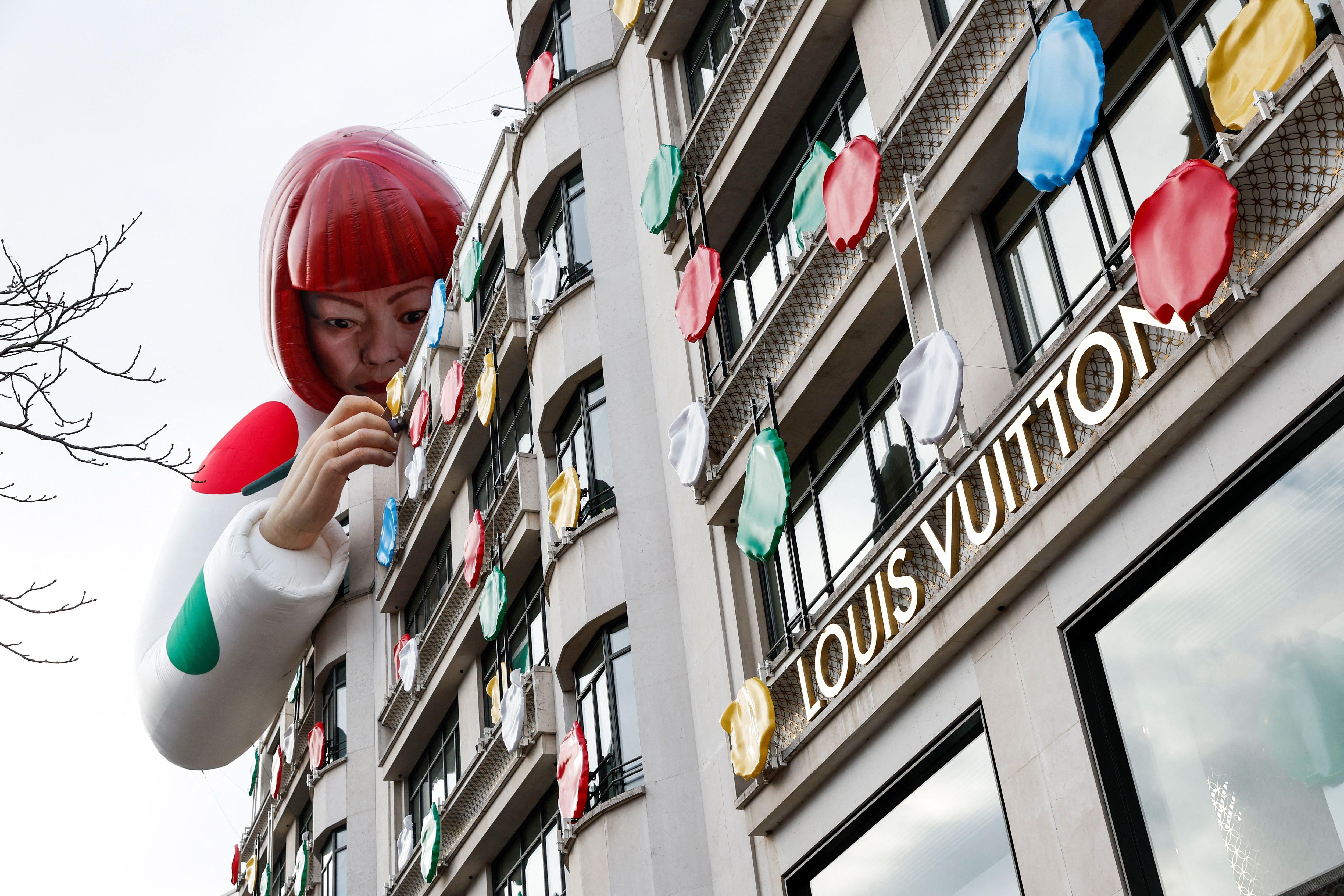 Exterior de la tienda Louis Vuitton con el logotipo y una figura artificial gigante de Yayoi Kusama inclinada sobre el tejado, aparentemente colocando lunares multicolores en el edificio.