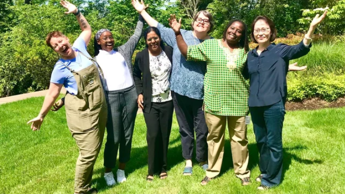 Un grupo de mujeres están de pie en fila sobre el césped, en lo que parece ser un jardín soleado. Todas sonríen y tienen las manos en alto. 