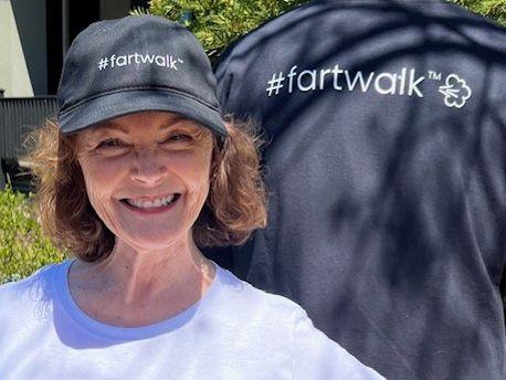 Mairlyn smiling at the camera, wearing a grey baseball cap with fartwalktm written on it. She has brown curly hair cut into a bob and is wearing a pale t-shirt. She is pictured in front of a man's back, he is wearing a t-shirt which also has fartwalk tm written on it. 