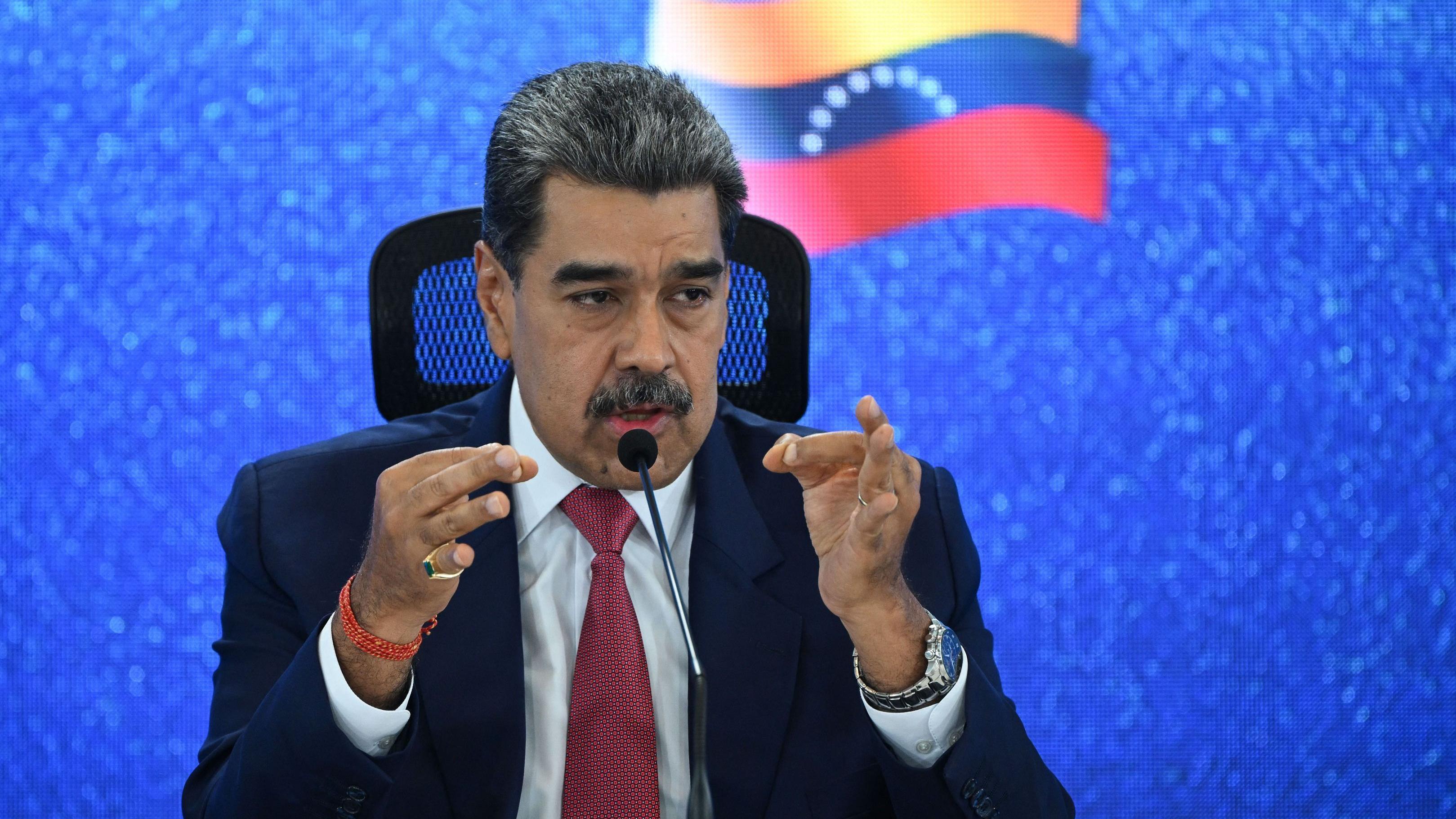 Nicolás Maduro habla frente a un micrófono y gesticula con las manos, mientras en el fondo se ve una pantalla led azul con una bandera de Venezuela.