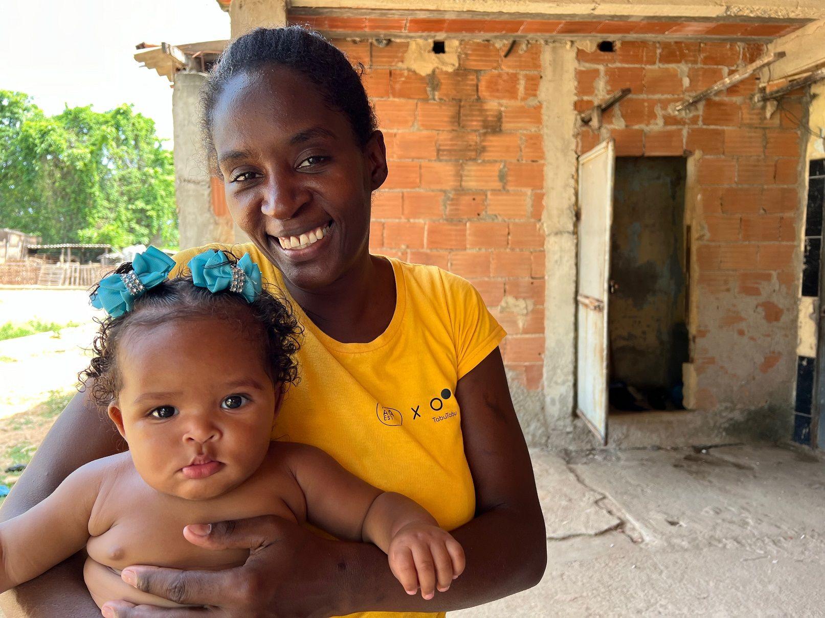 Evelyn sorrindo em pátio de casa, com bebê no colo