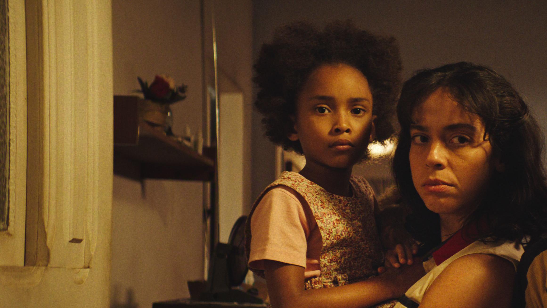 Uma mulher segura no colo uma crian&ccedil;a em cena de Emerg&ecirc;ncia Radioativa. Elas est&atilde;o em uma casa com ilumina&ccedil;&atilde;o quente, pr&oacute;ximas uma da outra, olhando em dire&ccedil;&atilde;o &agrave; c&acirc;mera com express&otilde;es s&eacute;rias e tensas; ao fundo, h&aacute; uma parede com uma pequena prateleira e objetos.