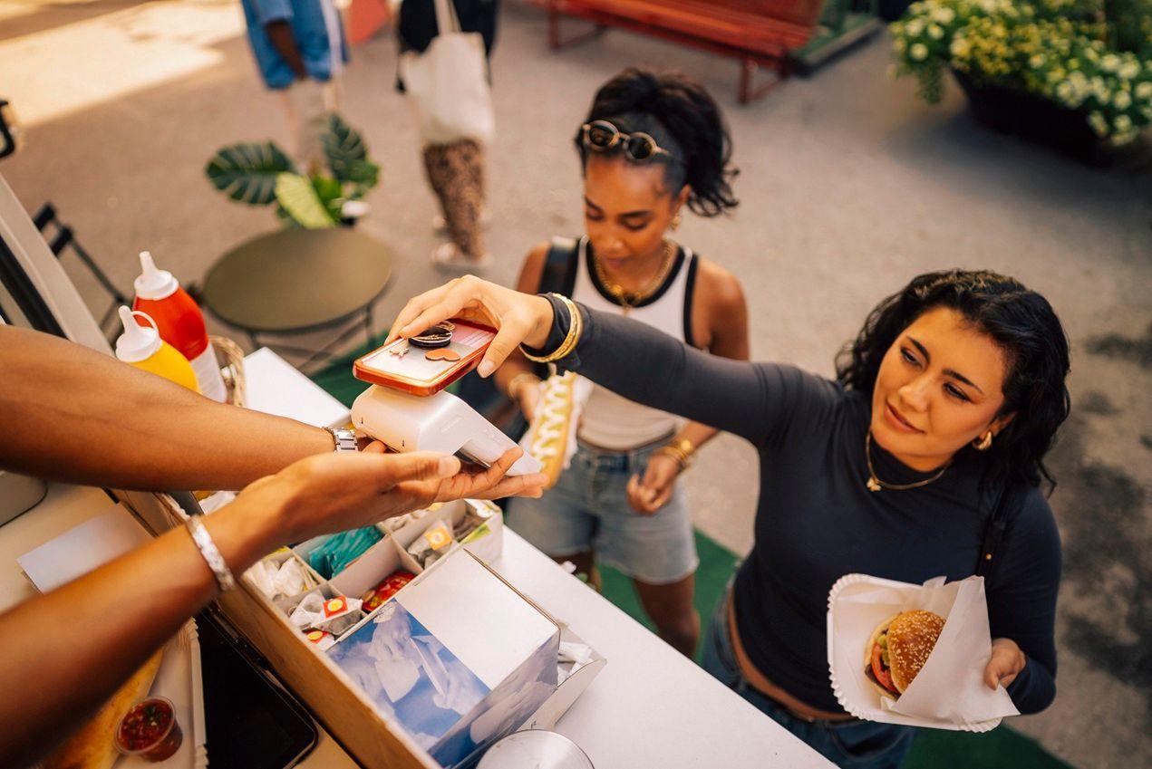Mulher est&aacute; pagando com celular por hamburguer que comprou em carrinho de comida. Ela aproxima o aparelho de uma maquininha de cart&atilde;o.