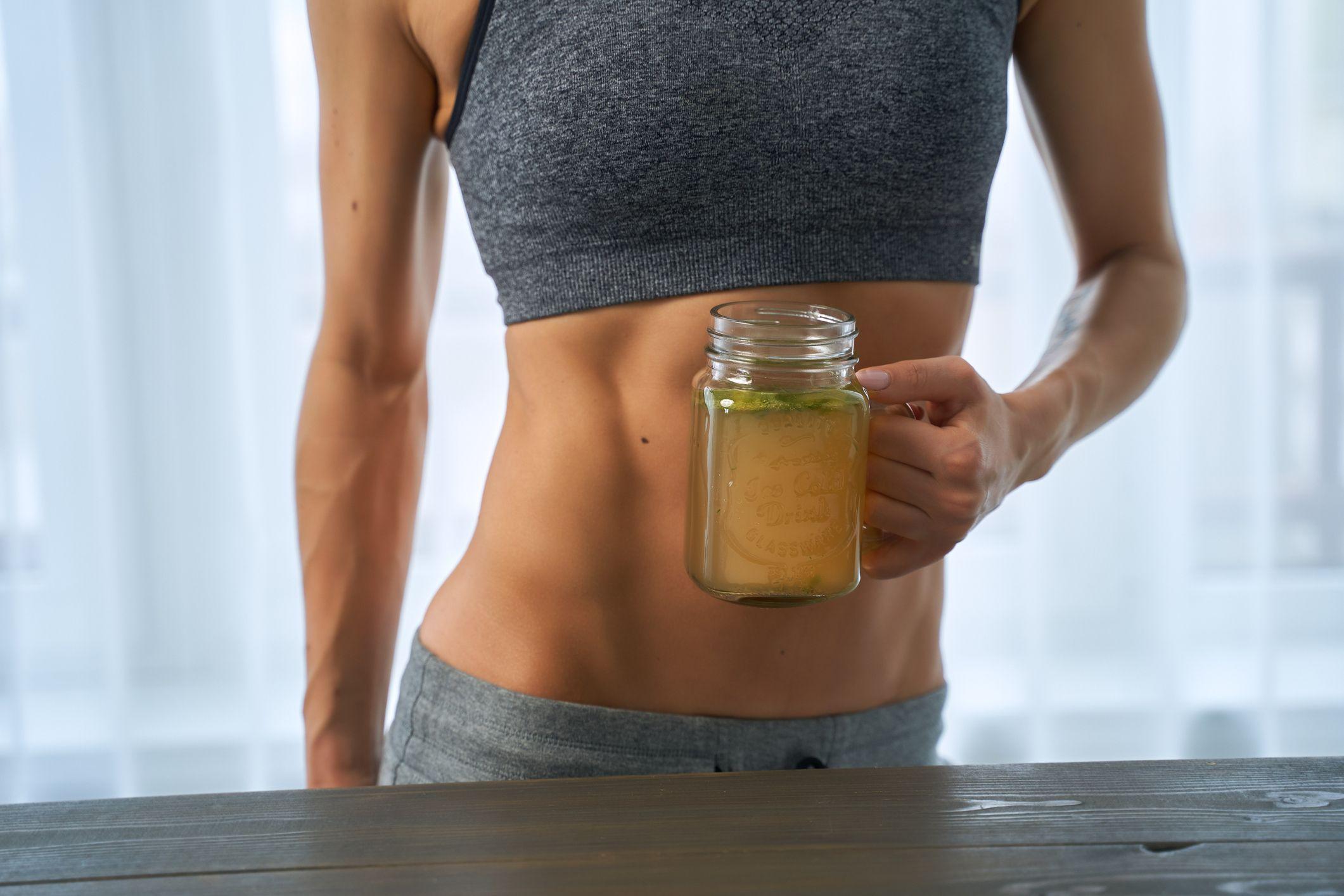Una mujer muy marcada, con ropa de ejercicio, sostiene una taza transparente con caldo de costilla.