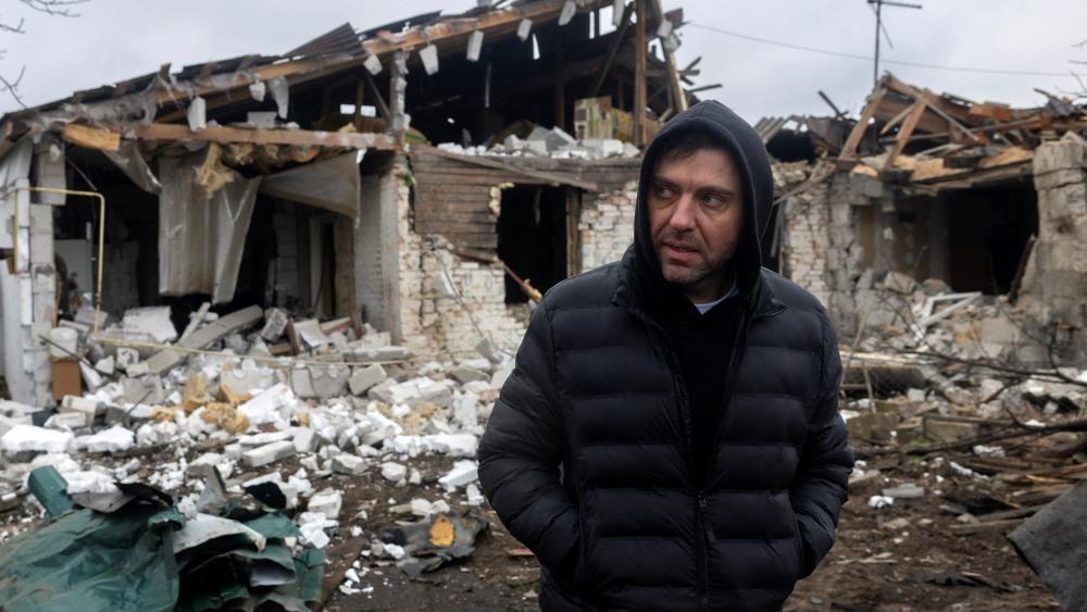 رجل يرتدي سترة منفّخة داكنة مع قلنسوة يقف أمام منزل تضرّر جرّاء هجوم، وقد تطاير سقفه وتناثرت الحجارة في كل مكان.