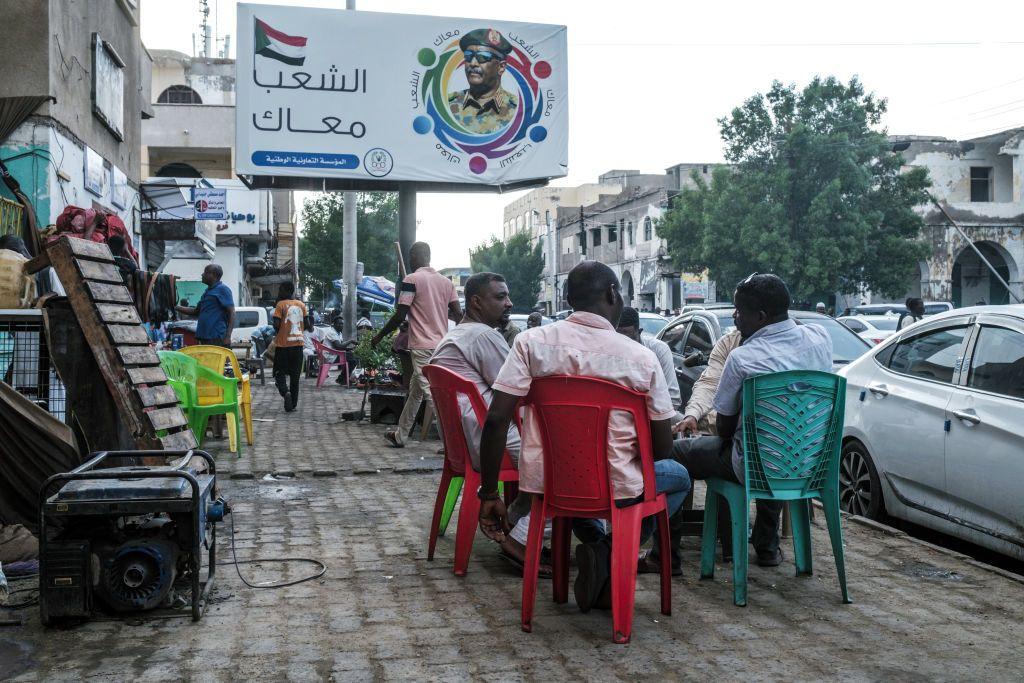 يجلس زبائن خارج مقهى بالقرب من لافتة تحمل صورة الفريق أول عبد الفتاح البرهان، القائد العسكري والحاكم الفعلي للسودان، في بورتسودان، مكتوب على اللافتة الشعب كله معاك.