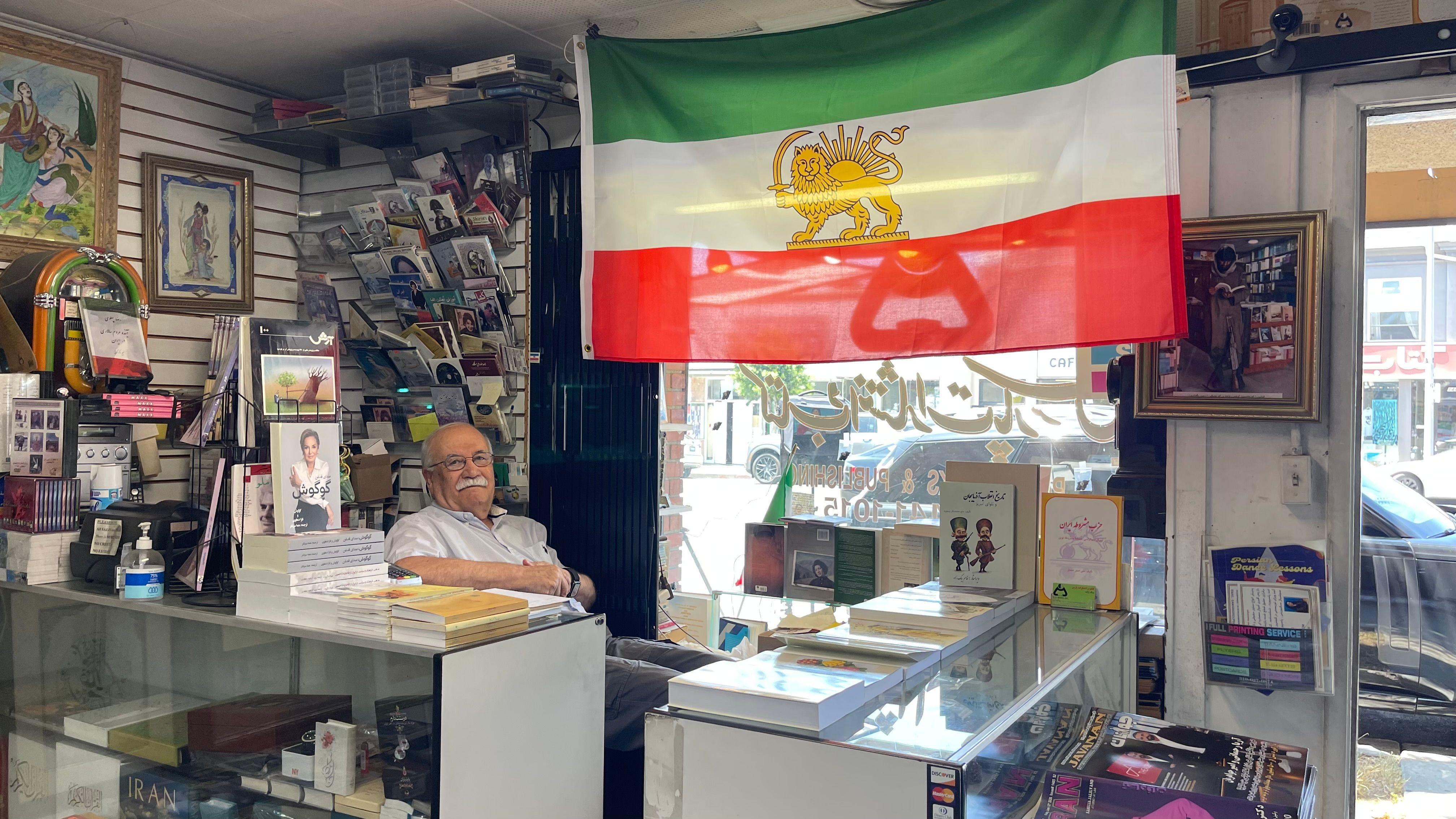 Un miembro de la diáspora iraní en Los Ángeles mira a cámara sentando detrás del mostrador de su librería, en la que tiene colgada la bandera previa a la revolución islámica con el león y el sol, en Teherángeles, Los Ángeles, Estados Unidos, el 15 de junio de 2026. (Foto: Leire Ventas)