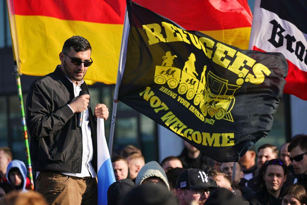 Simpatizantes en un acto de AfD junto a una bandera que afirma en inglés "Refugiados no bienvenidos".
