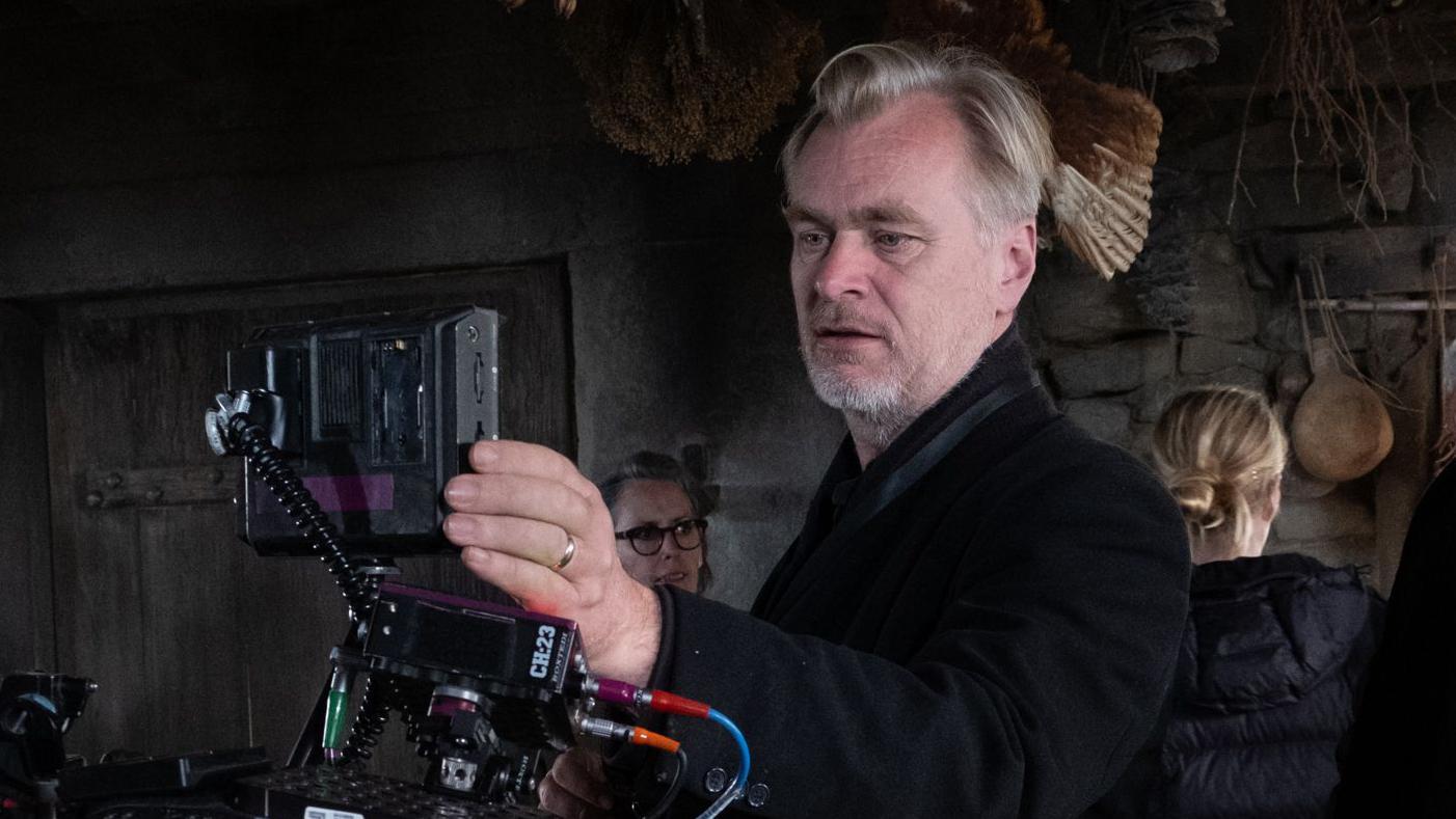 O diretor Christopher Nolan veste uma jaqueta preta e est&aacute; no centro da imagem, olhando atrav&eacute;s de uma c&acirc;mera IMAX