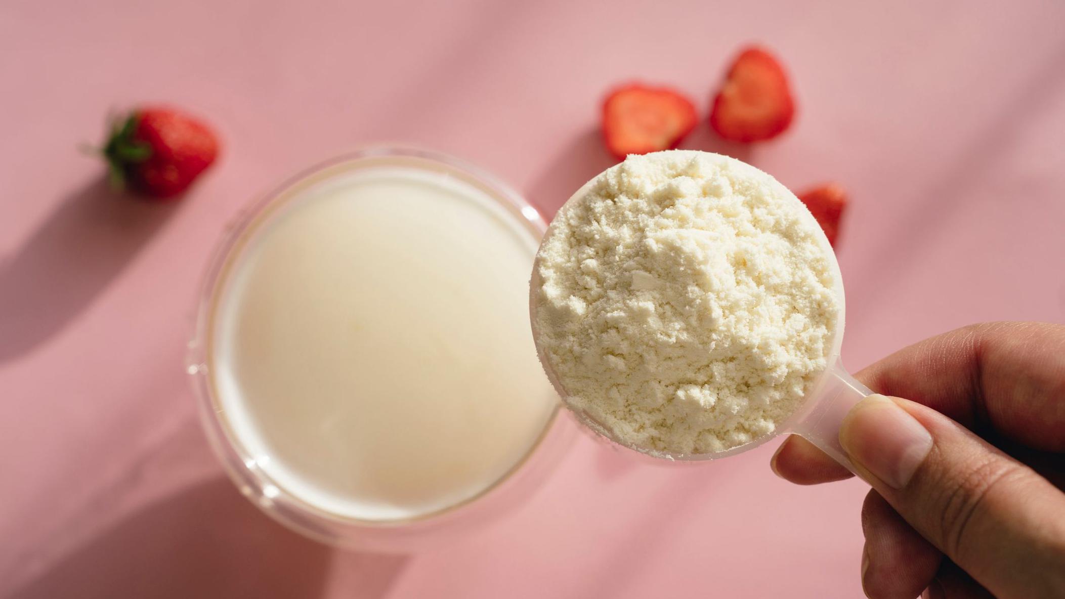 Una mano sostiene una cucharada de proteína en polvo sobre un vaso de leche. Al fondo hay un fondo rosa y fresas esparcidas.