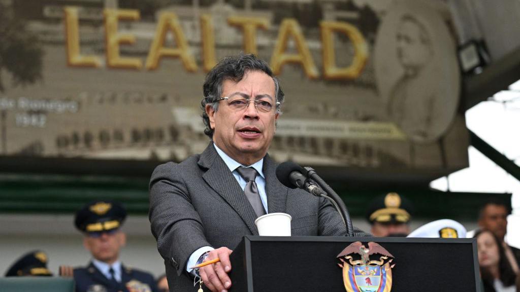Gustavo Petro de pie en un podio mientras le habla a varios militares. Viste un saco gris y una corbata del mismo color. Su camisa es azul. 