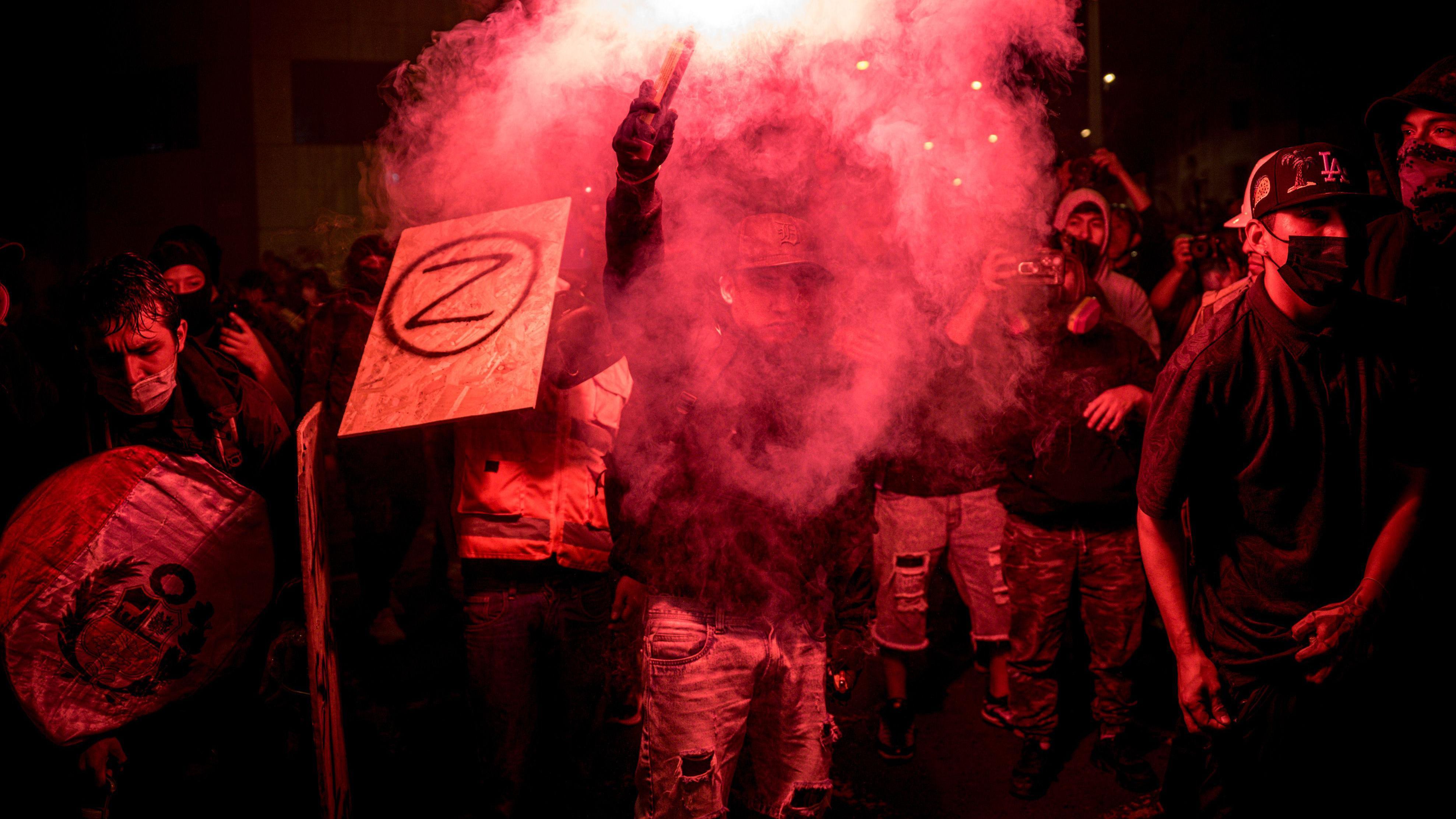 Un joven manifestante porta una bengala, rodeado por otros que exhiben pancartas con la letra "Z" y la bandera de Perú. 