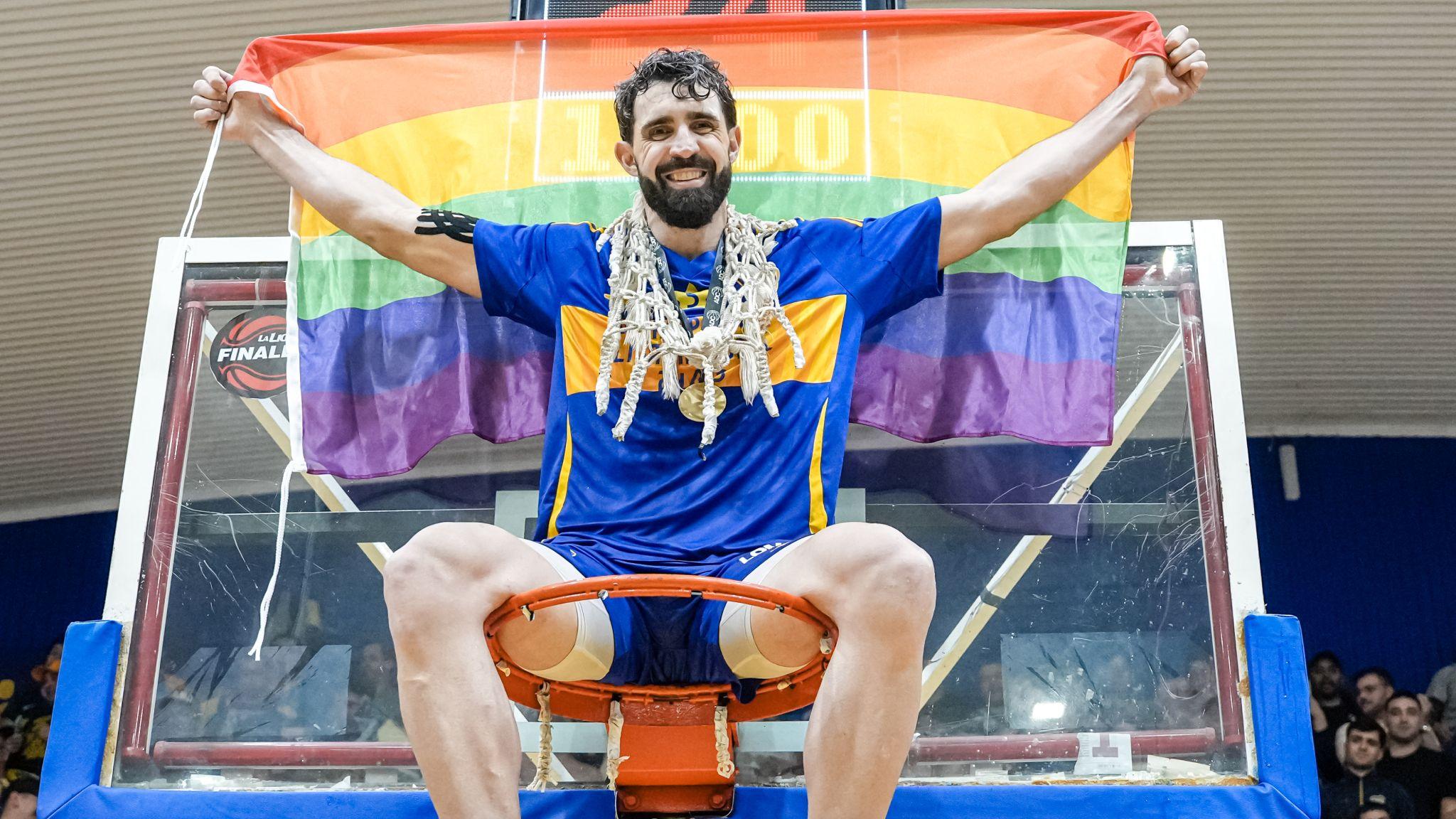 Sebastián Vega, sentado sobre uno de los aros de la cancha de básquetbol vistiendo la indumentaria de Boca Juniors, con la red del aro alrededor de su cuello y los brazos extendidos sosteniendo una bandera del orgullo gay detrás de su espalda.