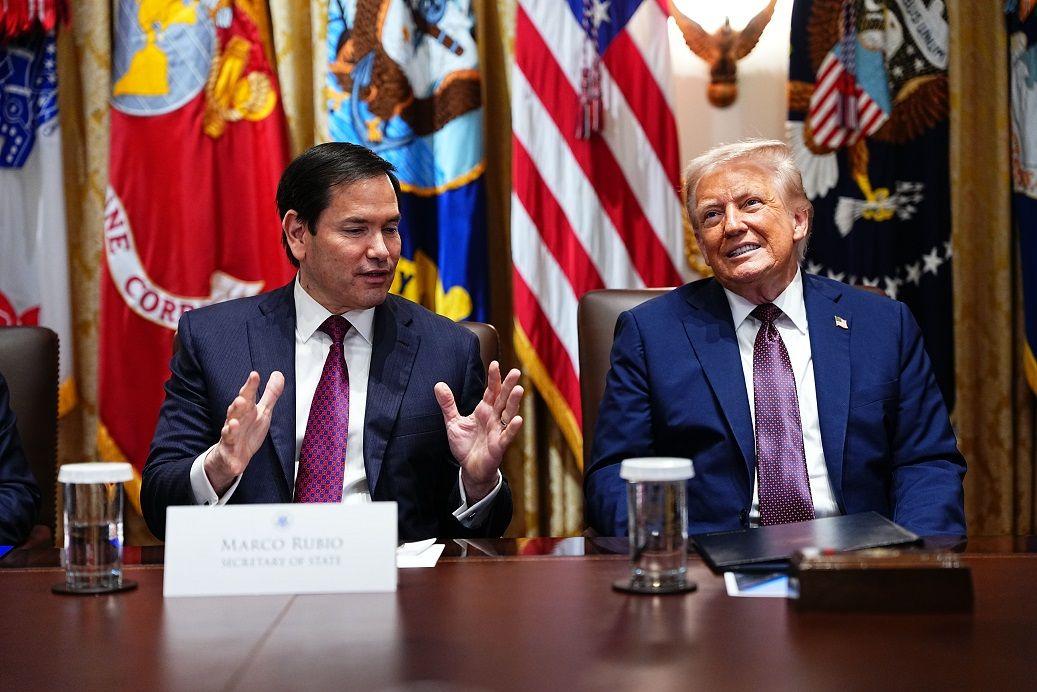 Rubio e Trump sentados diante de mesa, em frente a bandeiras; Rubio gesticula enquanto fala, e Trump sorri olhando para a frente