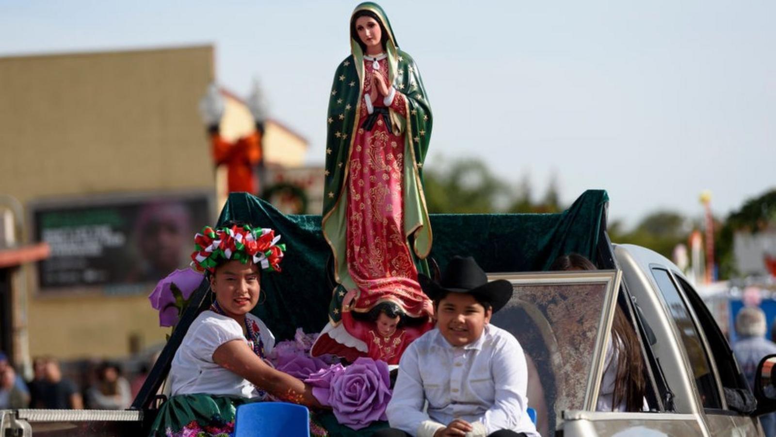 Una chica y un chico adolescentes de origen mexicano en Estados Unidos, sentados en el platón de una camioneta que carga una estatua de la Virgen de Guadalupe. La chica lleva una corona de rosas, el chico un sombrero de vaquero negro