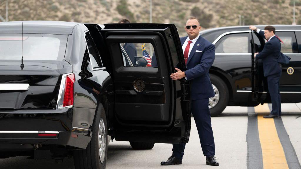 Dos agentes del Servicio Secreto de Estados Unidos, ambos de traje y uno de ellos con gafas de sol, mantienen abiertas las puertas traseras de dos versiones de la limusina blindada 