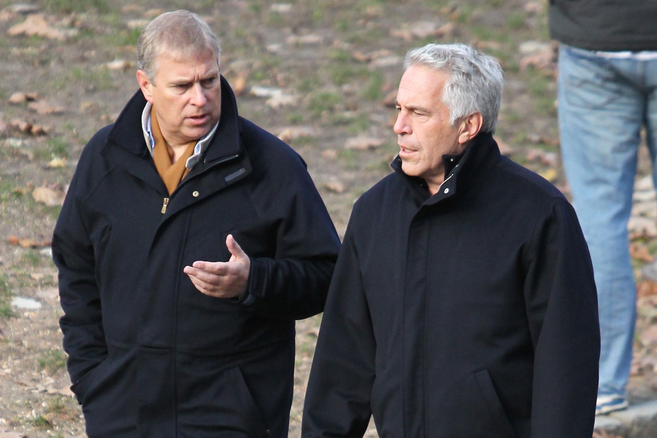 Andrew talks to Jeffrey Epstein as they walk. Both are wearing dark coats. Andrew's left hand is gesturing as he talks to Epstein. The ground behind them has grass and brown leaves on it. Someone with blue jeans is behind them and only their legs and waist are visible.