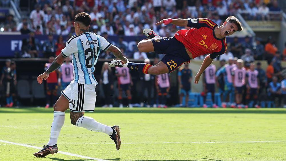 A imagem mostra uma disputa de bola durante uma partida de futebol entre Argentina e Espanha. Um jogador da sele&ccedil;&atilde;o espanhola aparece no ar ap&oacute;s um choque com um advers&aacute;rio argentino, enquanto o est&aacute;dio lotado e os bancos de reservas ao fundo refor&ccedil;am a intensidade do momento capturado durante o jogo.