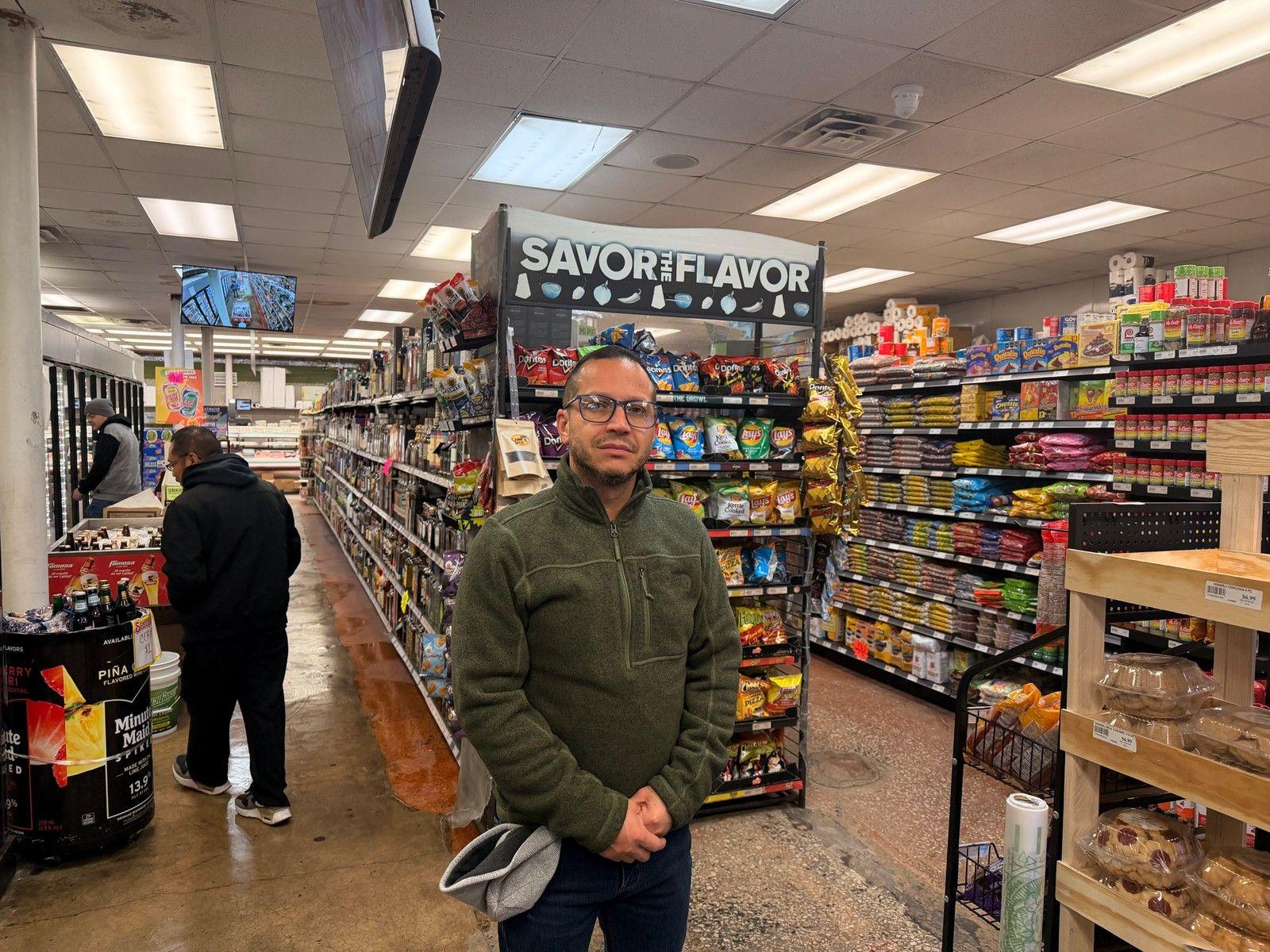 Carlos Macias parado frente a los estantes de su pequeña tienda de comestibles. Tiene anteojos y viste una chaqueta verde oliva. Un cliente camina por una de las hileras del almacén