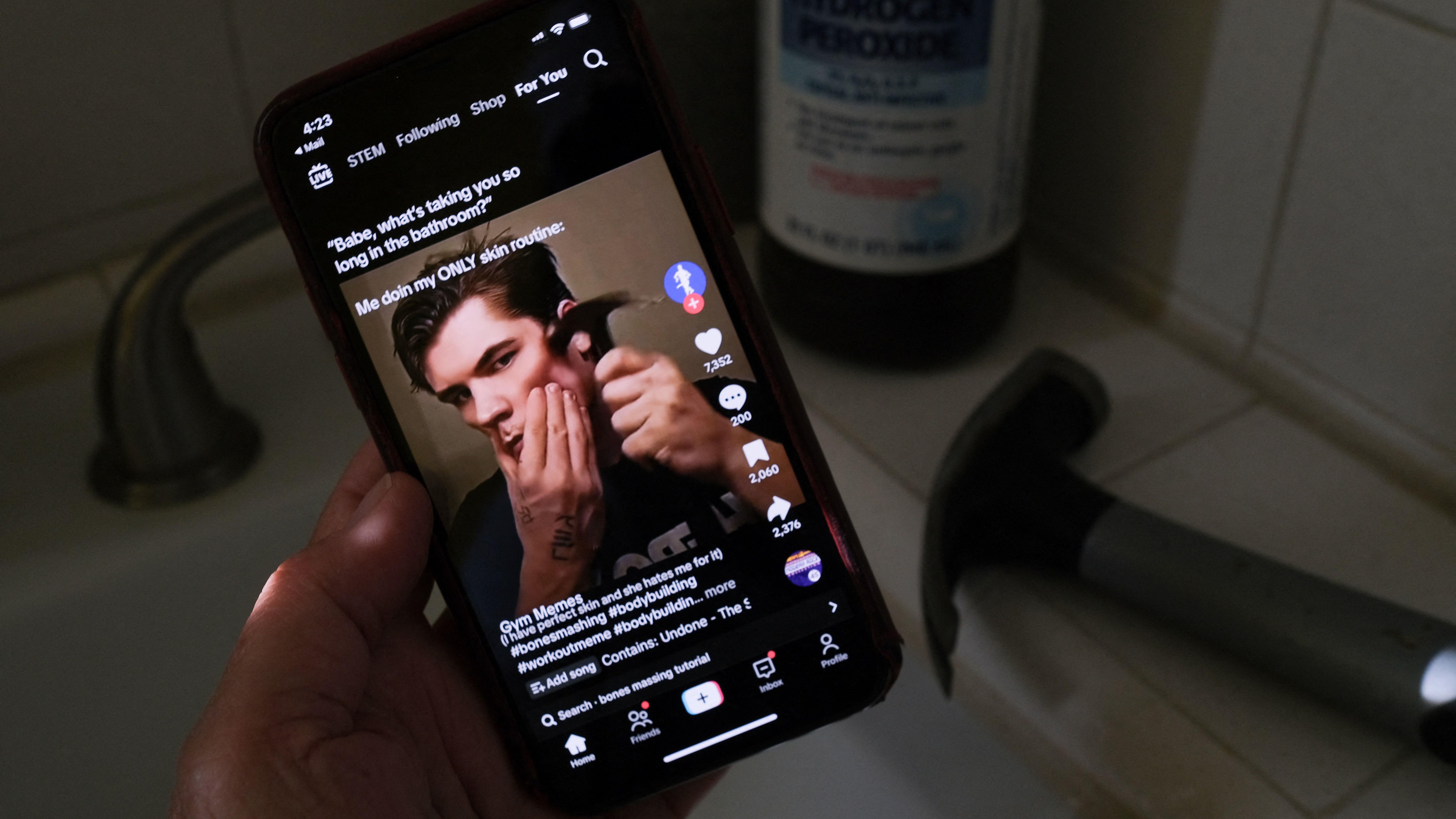 Um homem segura um smartphone exibindo um v&iacute;deo mostrando um jovem diante da c&acirc;mera em um conte&uacute;do relacionado a rotina de cuidados pessoais. A cena ocorre em um banheiro, identificado pela pia, torneira e pelos objetos ao redor, incluindo um frasco ao fundo e um aparelho el&eacute;trico sobre a bancada. A ilumina&ccedil;&atilde;o &eacute; baixa e concentra a aten&ccedil;&atilde;o na tela do celular.