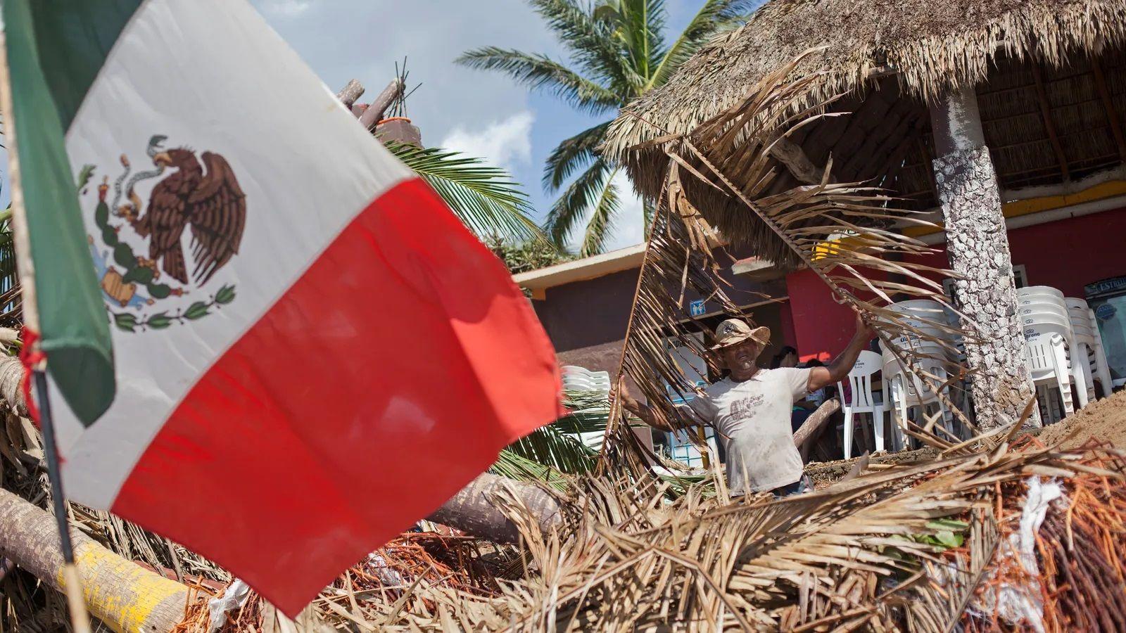 Una imagen muestra la bandera de México en medio de los daños causados por el huracán Patricia. Al fondo se ve un hombre con sombrebro limpiando los escombros.