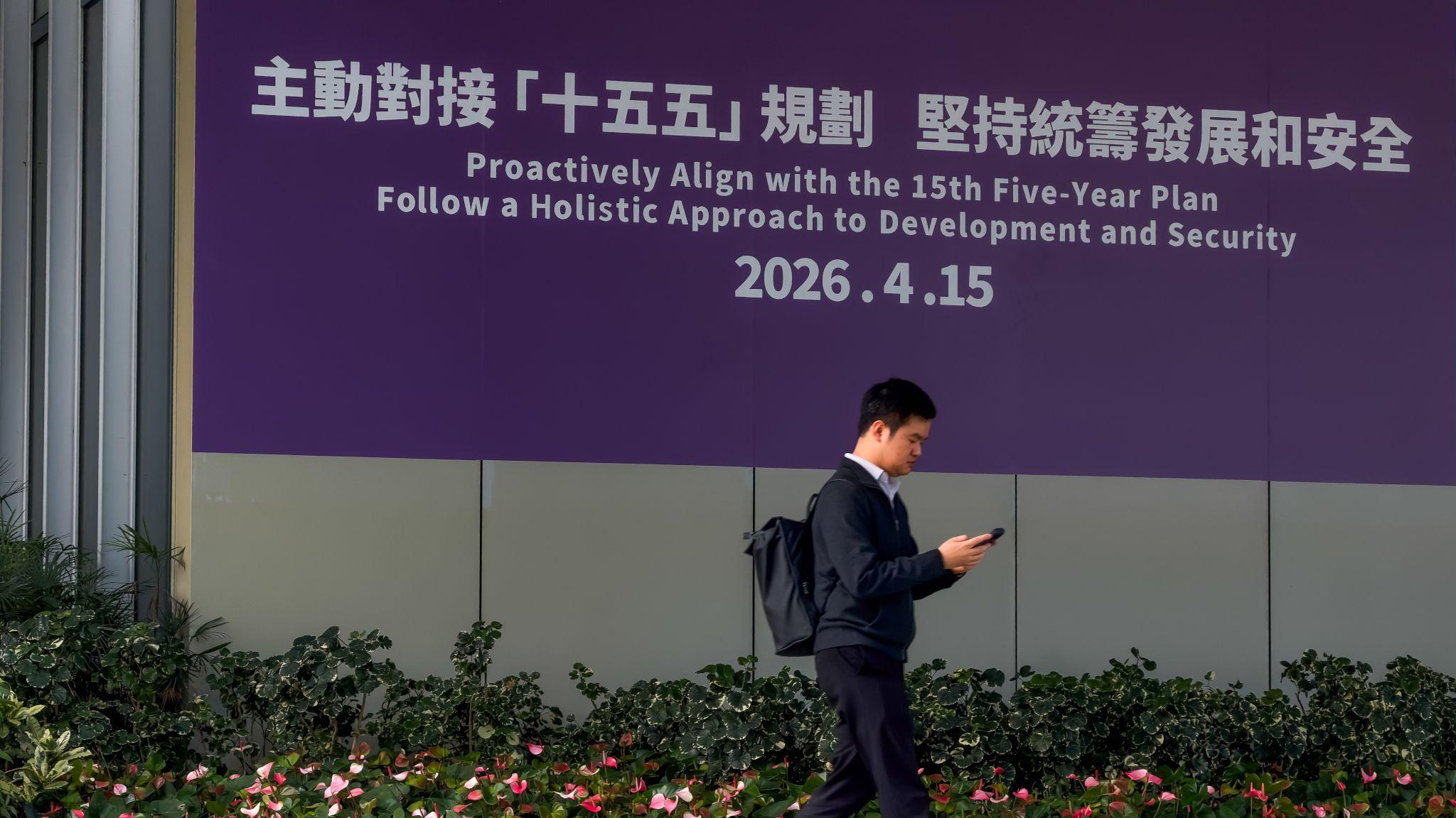 金鐘添馬中國香港特區政府總部外一位男子一邊查看手機一邊走過一面大型政治宣傳橫幅，上面印上標語「主動對接『十五五』規劃，堅持統籌發展和安全」（17/3/2026）