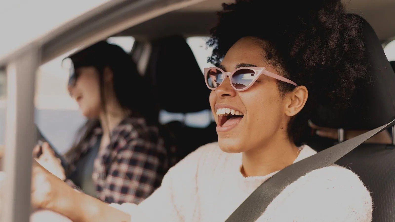صورة لامرأة ذات شعر أسود كثيف، ترتدي نظارات ويفتح فمها على اتساعه. تجلس في سيارة خلف المقود،