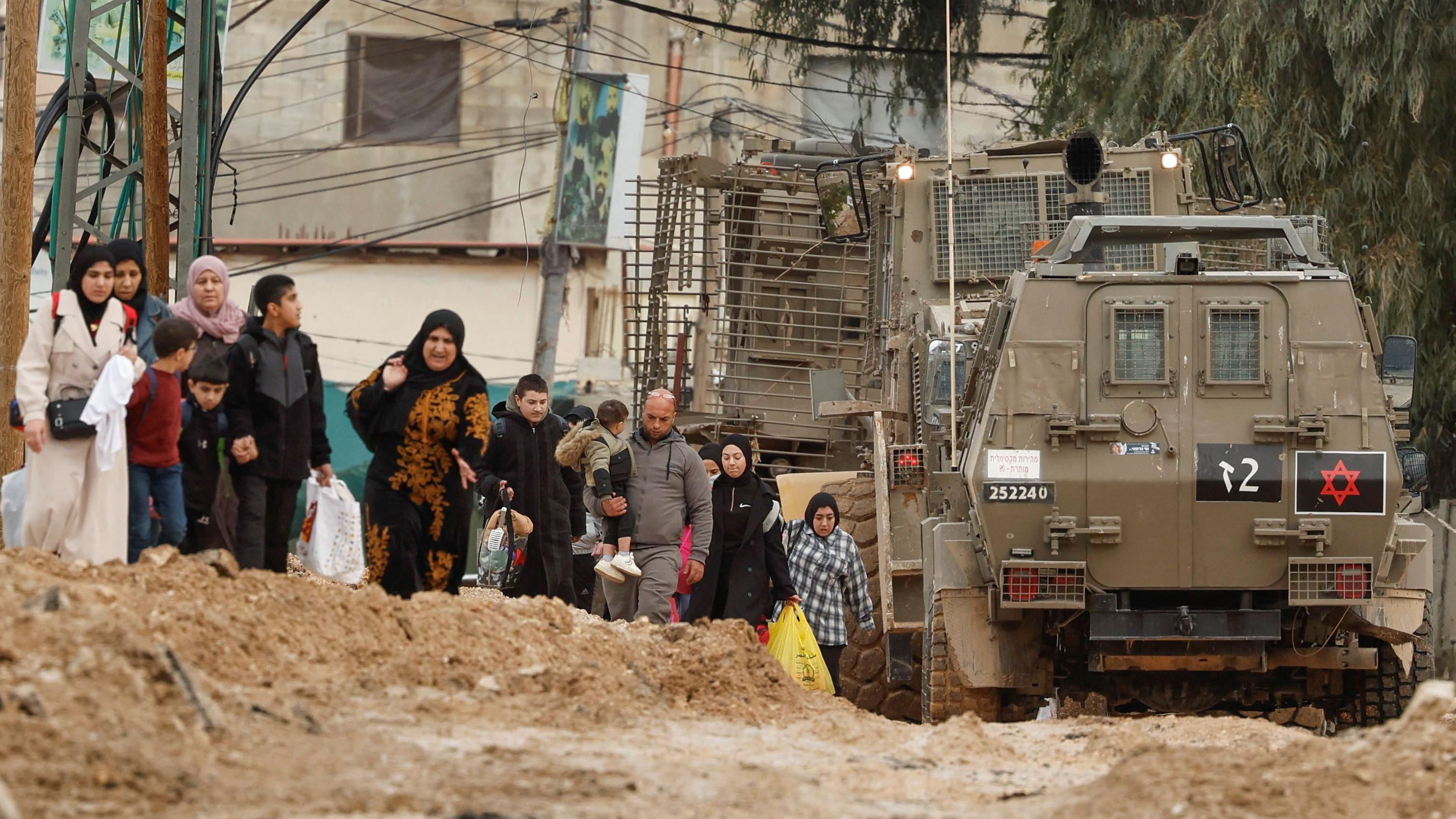 فلسطينيون يسيرون بجوار مدرعات إسرائيلية أثناء مغادرتهم مخيم جنين في الضفة الغربية التي تحتلها إسرائيل، في 22 يناير/كانون الثاني 2025