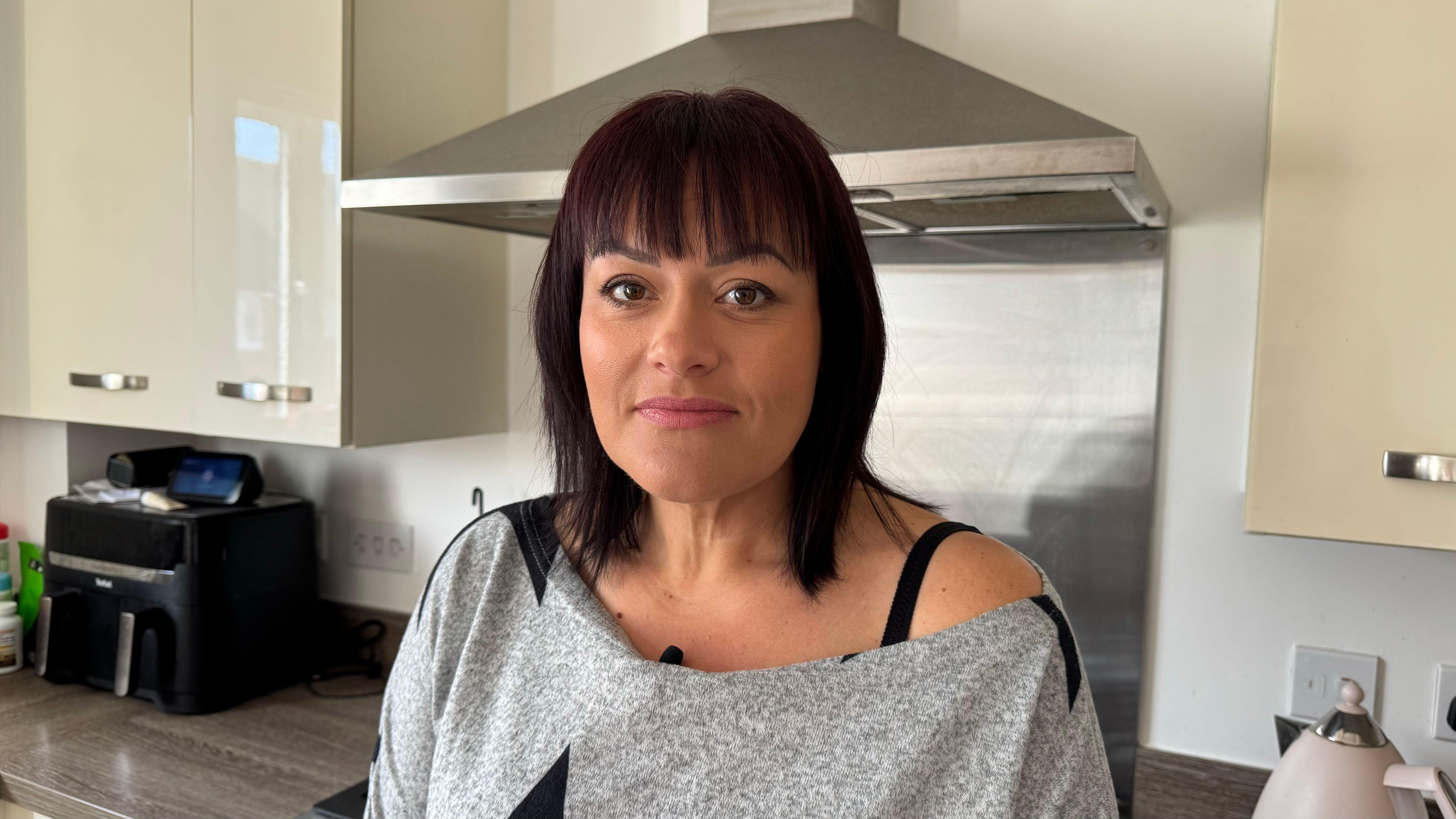 Hayley Dawe vestindo camiseta cinza com sua cozinha ao fundo, ela tem cabelo liso escuro com franja e olhos castanhos