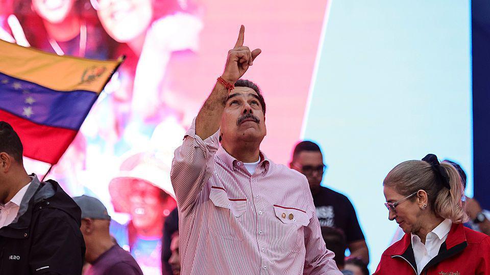 委內瑞拉總統尼古拉斯·馬杜羅身穿有領襯衫，袖子捲起，內搭白色T恤，站在巨型螢幕前，指向天空