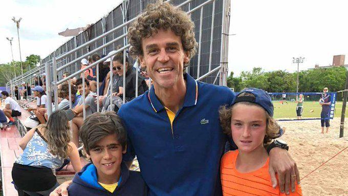 O tenista João Fonseca quando criança, vestindo camiseta laranja e boné azul ao contrário, ao lado do tenista Gustavo Kuerten e de uma segunda criança não identificada.