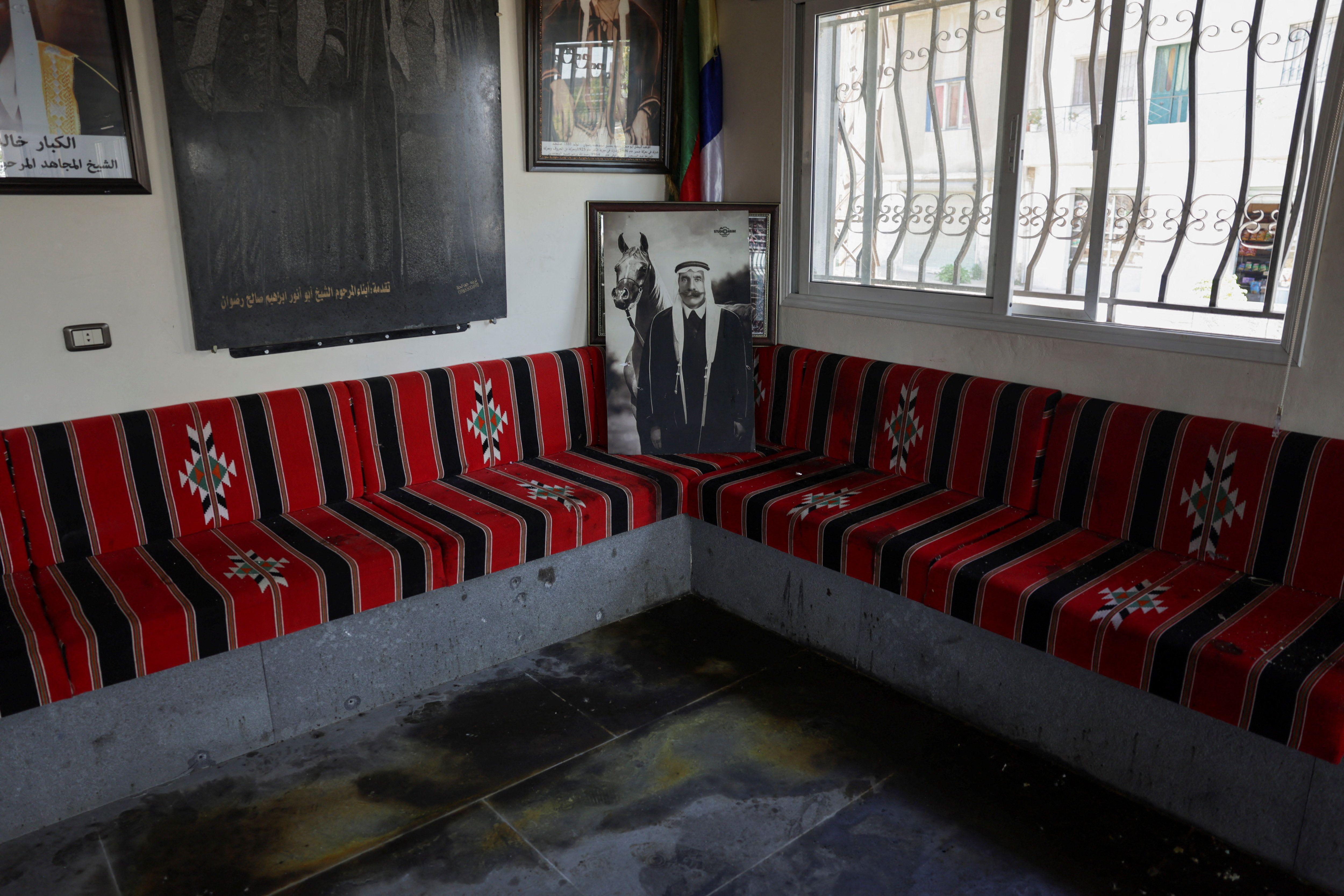 Un salón con todas las paredes jalonadas por bancos hechos de concreto, pero decorados por encima con una tela continua negra y roja, con motivos árabes. Hay una venta a la derecha, la bandera drusa en el centro y un retrato antiguo, además de otros retratos en la pared izquierda. El suelo está lleno de manchas. 