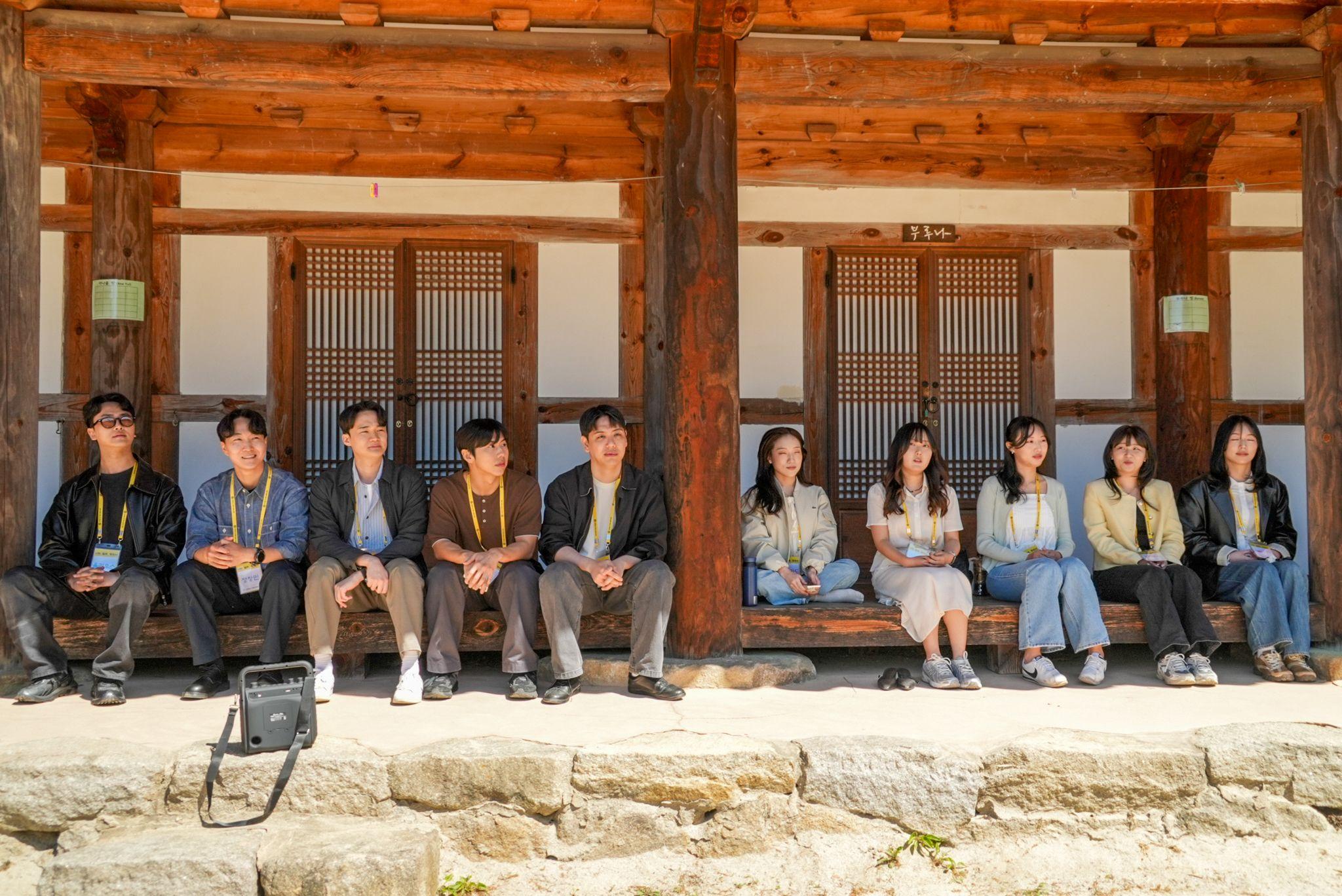 명찰을 착용한 남성 5명과 여성 5명이 대형 나무 기둥과 격자문이 있는 전통 한국 사찰의 목조 툇마루에 앉아 있다. 이들은 밝은 낮에 툇마루 곳곳에 흩어져 앉아 있으며, 앞쪽에는 돌로 된 기단과 가장자리 근처에는 가방 하나가 놓여 있다.