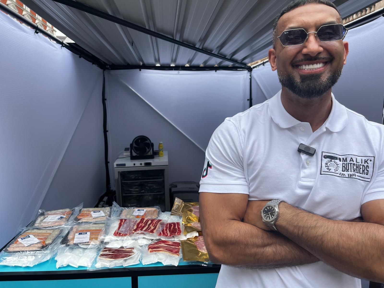 Manny Malik to the right of the shot wearing a white polo shirt with black Malik Butchers branding. To the left of the shot behind him is a selection of packed meats