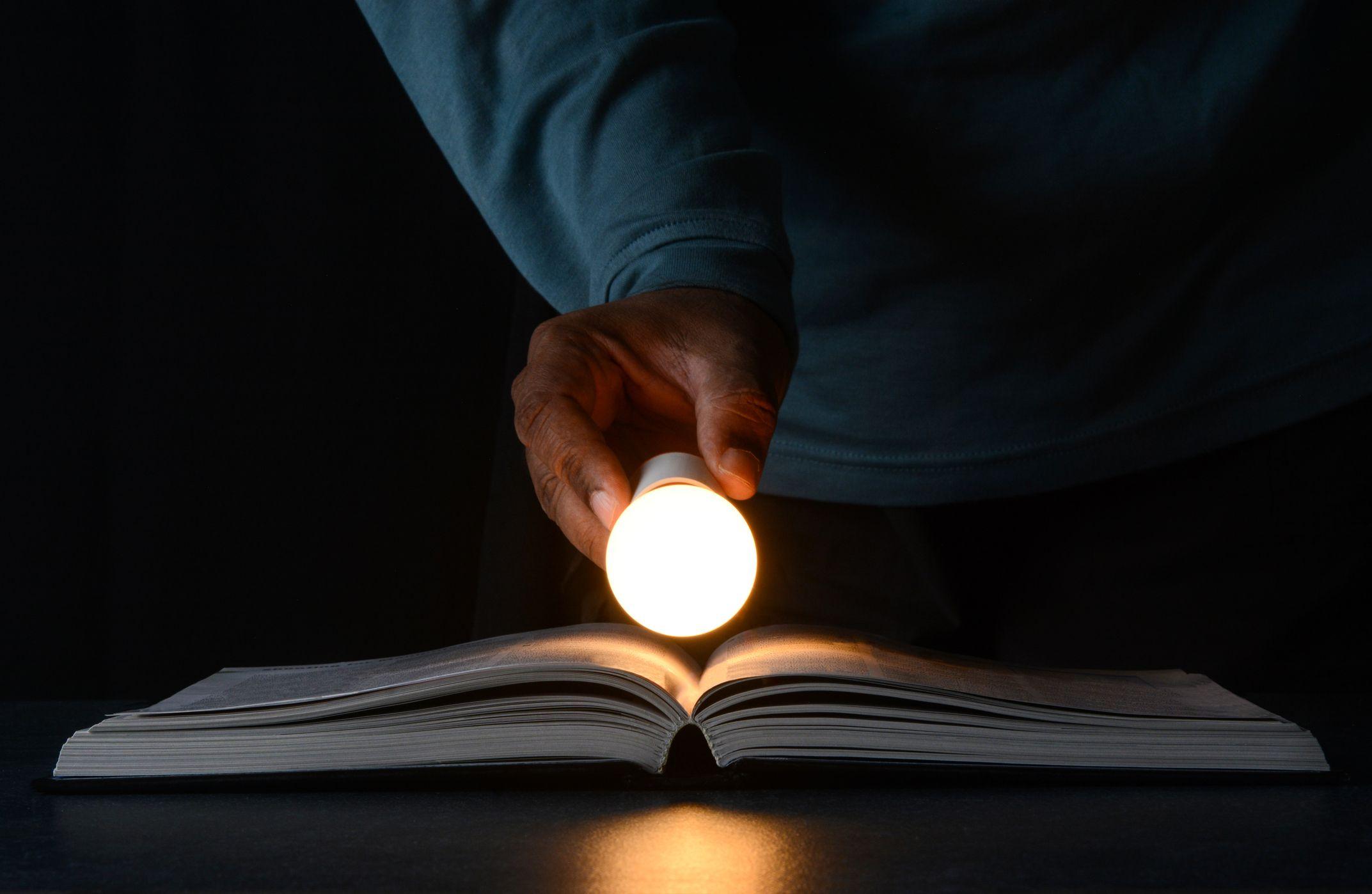 مفهوم الأفكار الإبداعية والتعلم والابتكار. رجل يحمل مصباحاً مضيئاً على كتاب مفتوح. تظهر فقط يد الرجل التي تبدو سمراء كما يبدو أنه يرتدي فميصاً أزرق اللون، مع المصباح والكتاب.