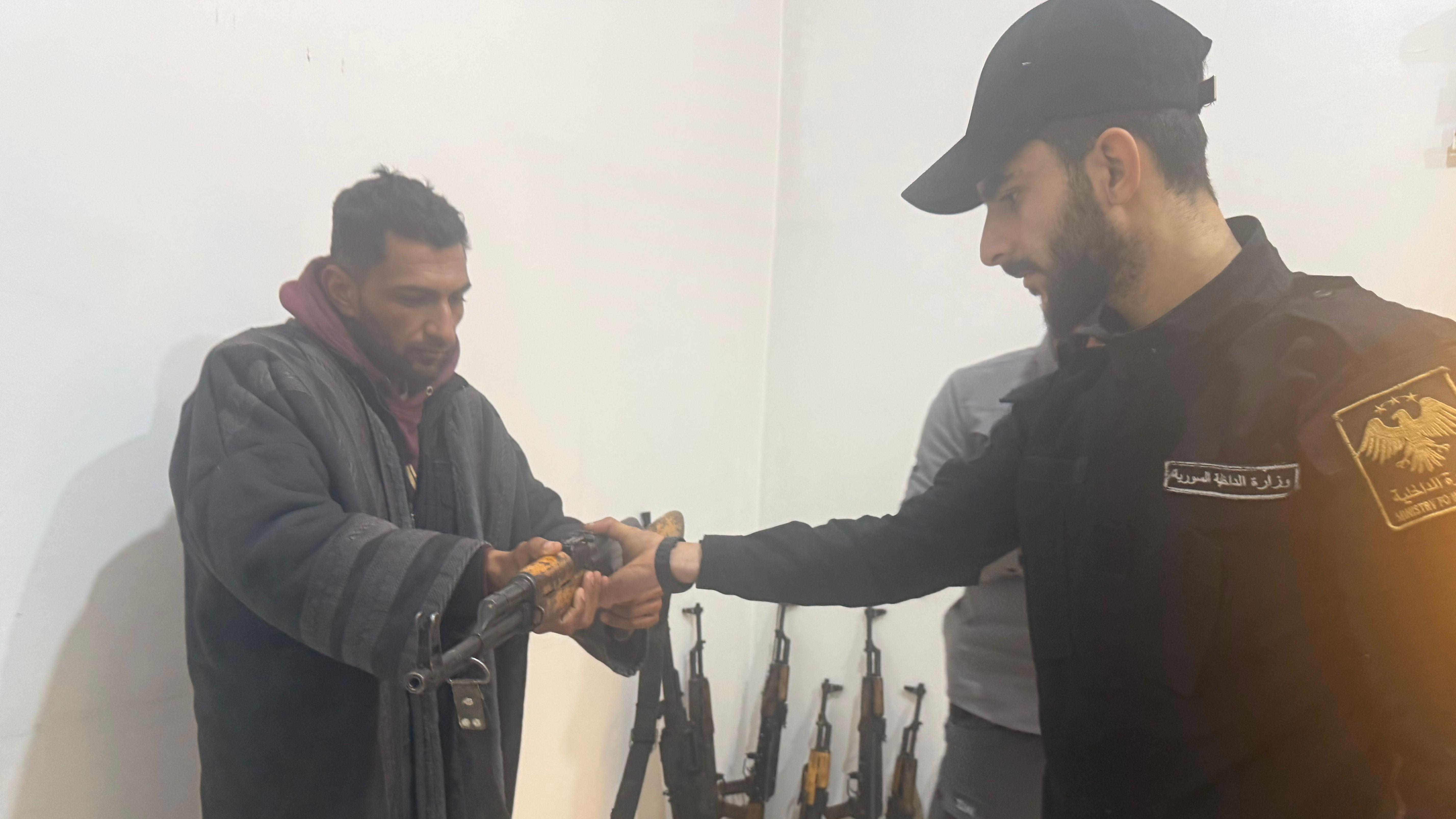رجل يرتدي معطفاً يسلم سلاحاً بيديه لرجل يرتدي زياً رسمياً مكتوب عليه "وزارة الداخلية السورية" 