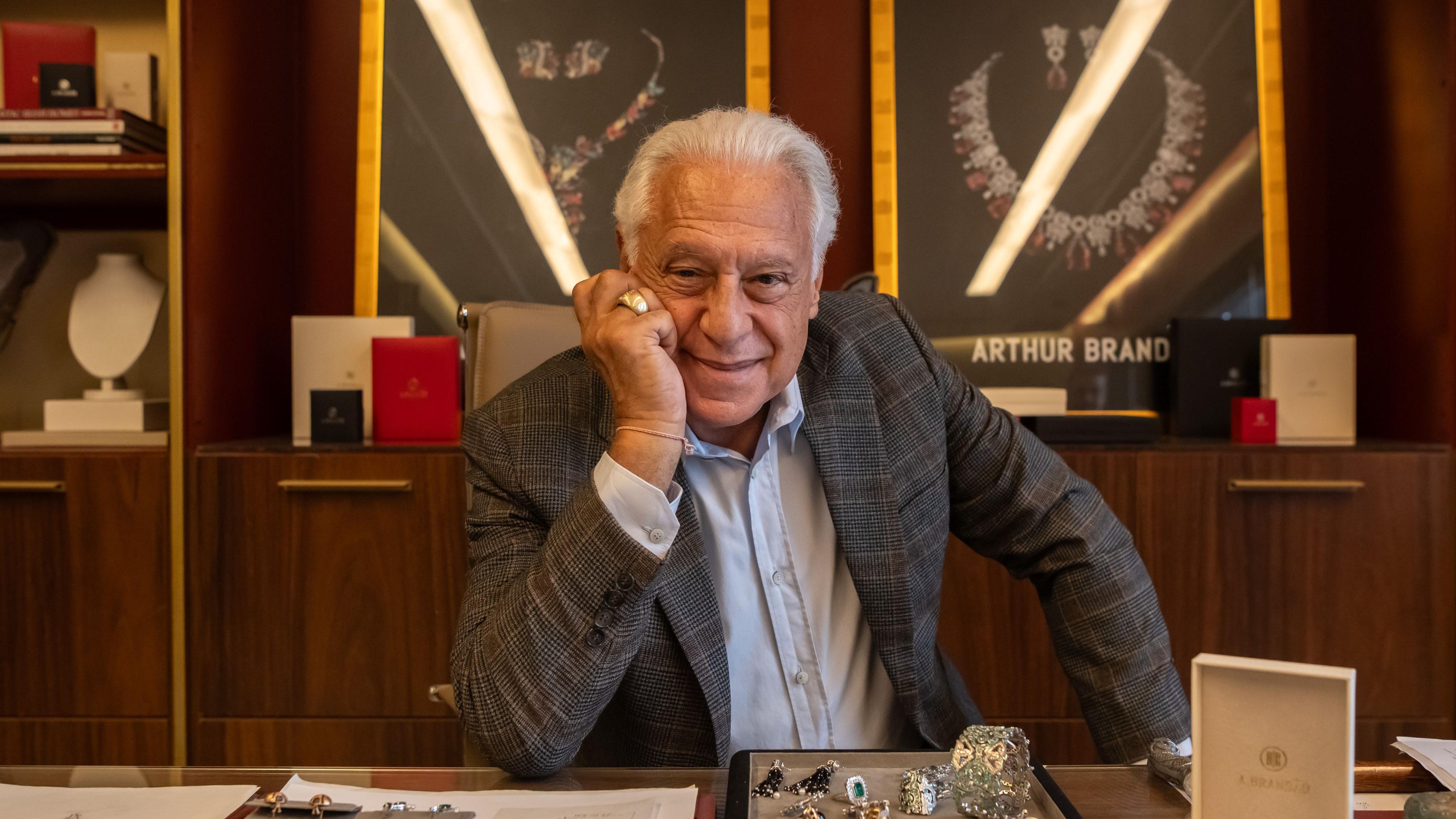 Antonio Fagundes, um homem de cabelos brancos, posa apoiando o rosto na m&atilde;o atr&aacute;s de uma mesa com joias e documentos, em um ambiente sofisticado de escrit&oacute;rio. Vestindo blazer xadrez e camisa clara, ele sorri para a c&acirc;mera enquanto, ao fundo, vitrines iluminadas exibem colares de cristais.
