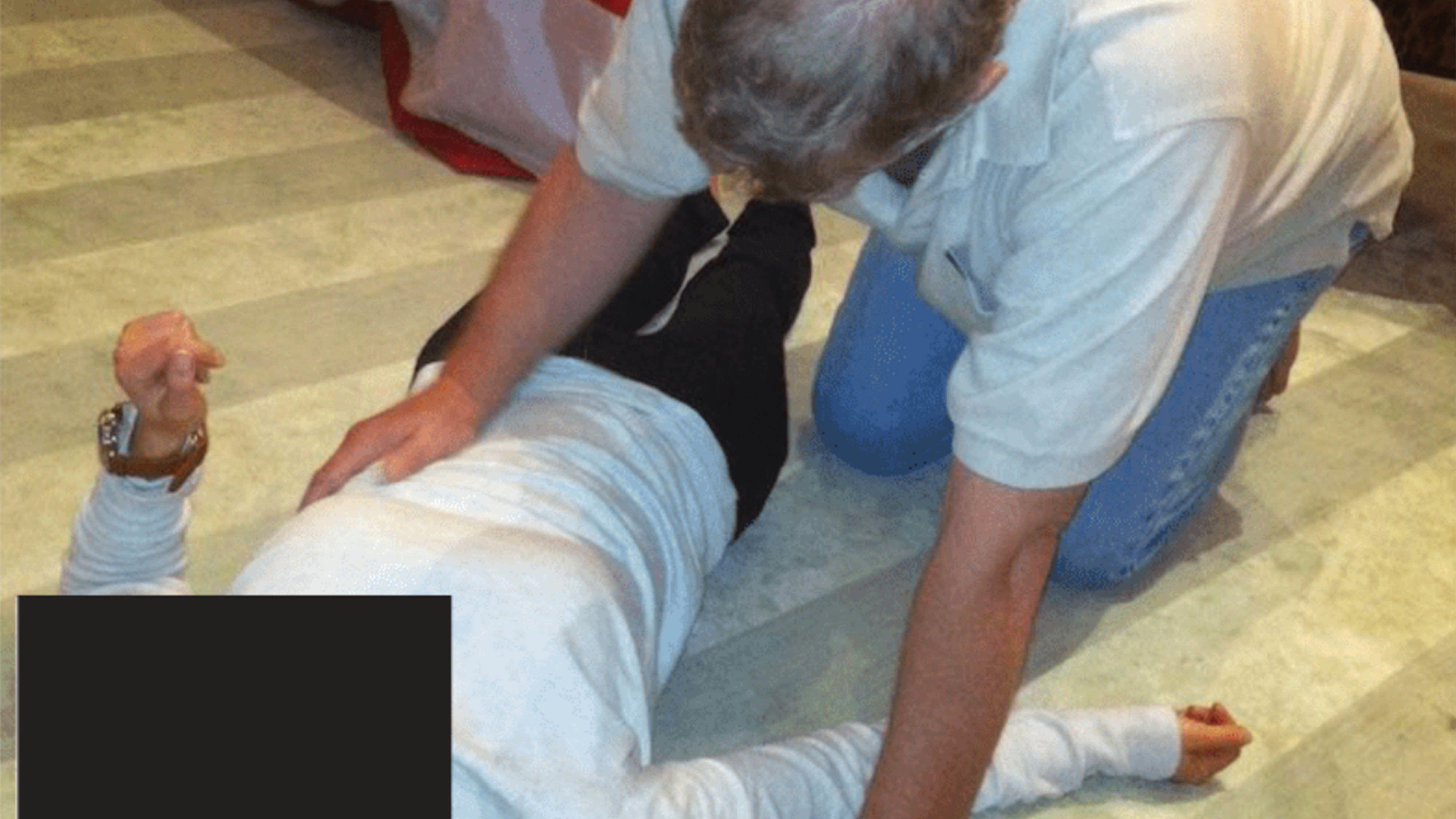 Um homem ajoelhado sobre uma mulher deitada no ch&atilde;o. O rosto dela est&aacute; oculto por um ret&acirc;ngulo preto. Ela veste cal&ccedil;as pretas e uma blusa branca e levantou o bra&ccedil;o esquerdo. O homem est&aacute; tocando a lateral do abd&ocirc;men dela.