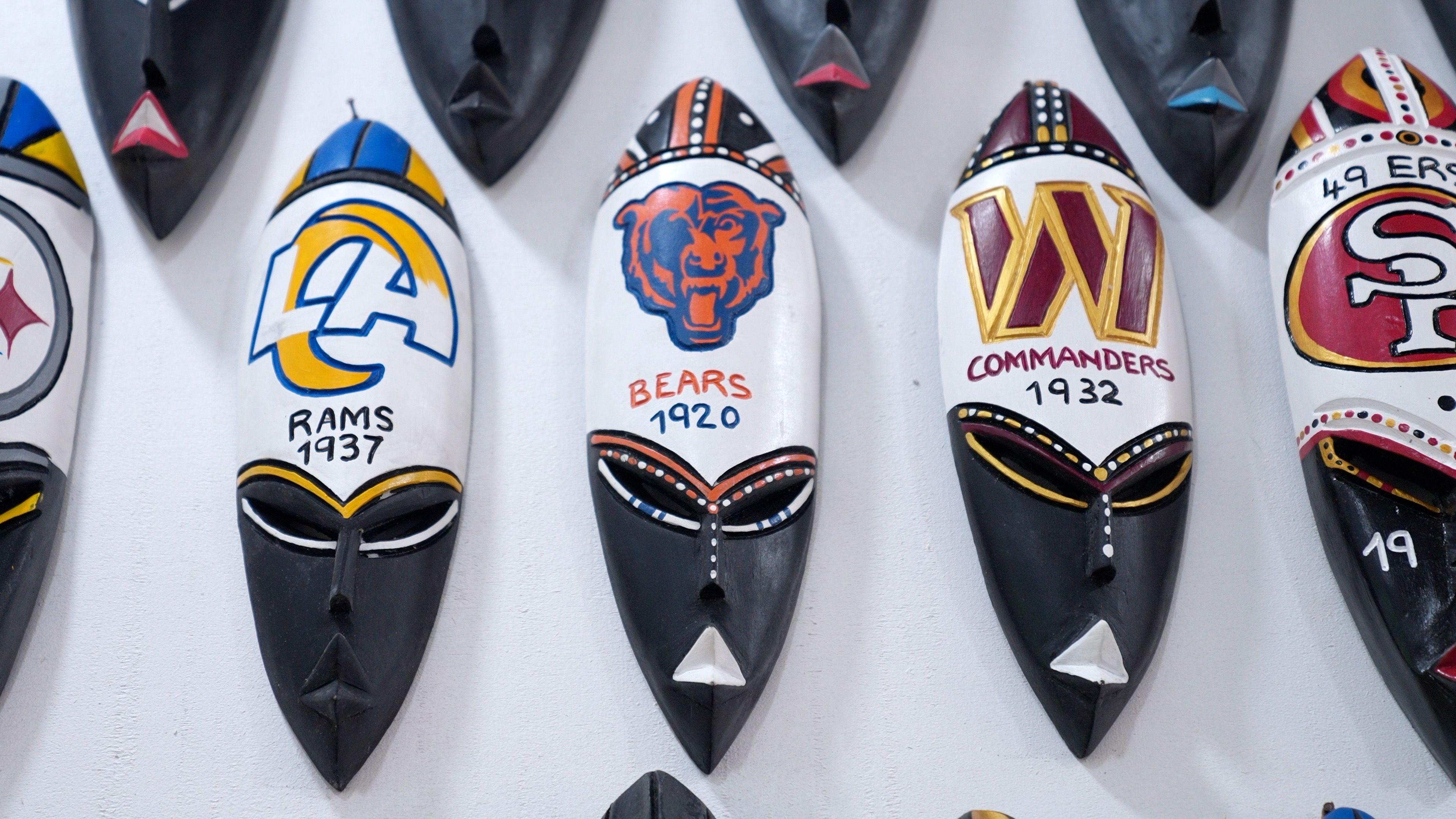 Una hilera de máscaras tradicionales ghanesas con los logotipos y nombres de equipos de fútbol americano: los Rams, los Bears y los Commanders, pintados en la parte superior