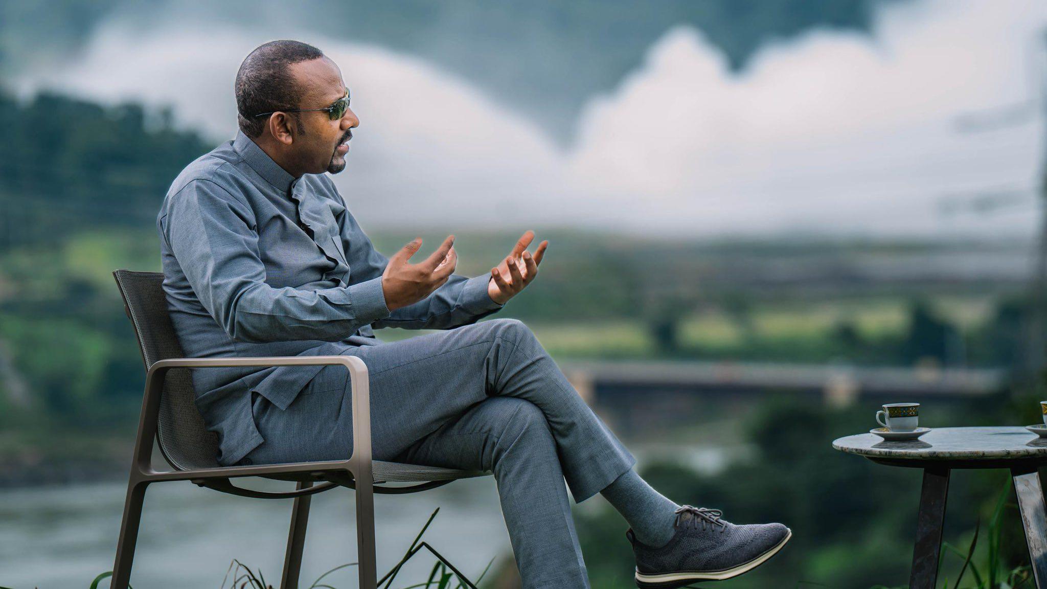 Abiy Ahmed in grey trousers and shirt sits on a chair with a coffee table in front of him. He is holding his hands out to make a point to an unseen interviewer. Behind him, blurred, are the gushing waters of the Blue Nile. 