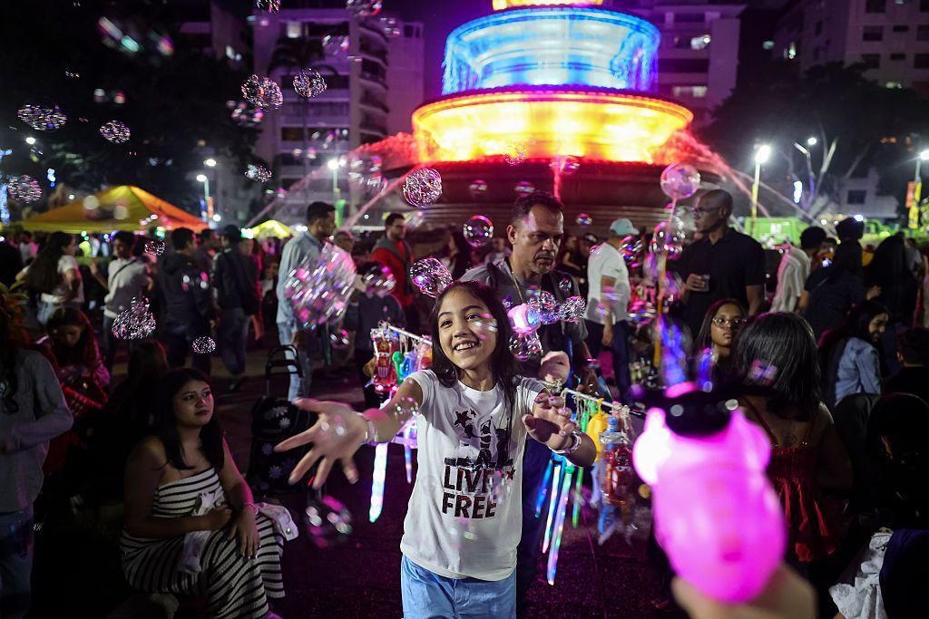 Una niña juega con burbujas de jabón que flotan en el aire frente a una fuente iluminada de colores, rodeada de muchas personas más