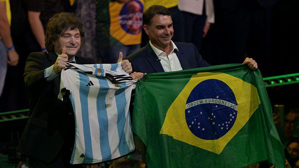 Javier Milei muestra la camiseta de la selección argentina de fútbol durante un acto en Sao Paulo en julio para lanzar la candidatura presidencial de Flávio Bolsonaro, quien exhibe a su lado la bandera de Brasil