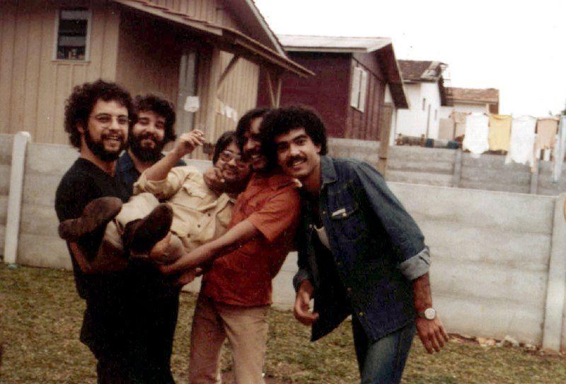 A imagem mostra cinco homens posando juntos ao ar livre em frente a casas simples e um muro baixo. Dois deles seguram outro homem no colo enquanto todos sorriem para a c&acirc;mera. A foto tem apar&ecirc;ncia antiga, com tons amarelados, e ao fundo h&aacute; roupas penduradas em um varal.