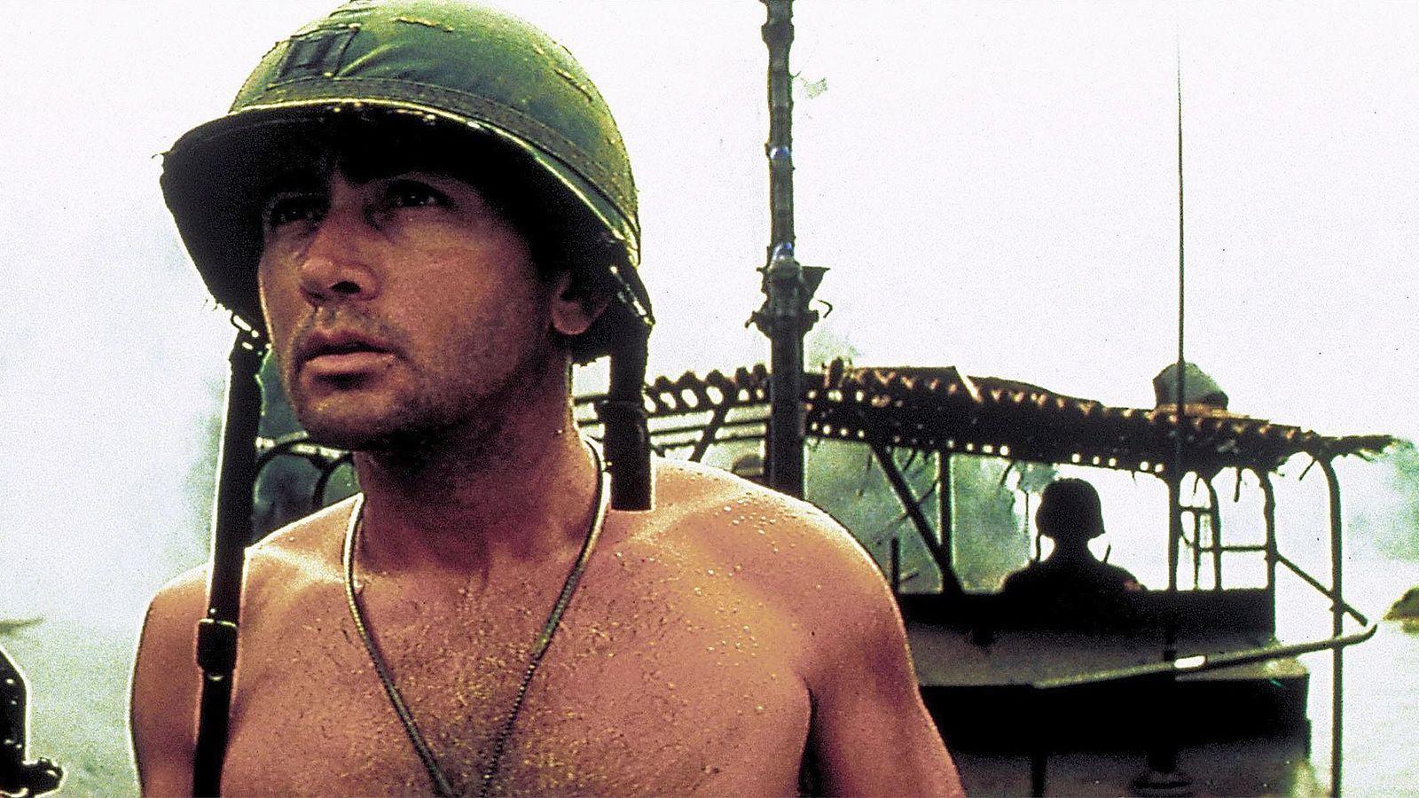 Martin Sheen, representanto a un oficial del ejército de EE.UU. con un casco, sin camisa, con un barco en el fondo, en una escena de "Apocalipsis Now"