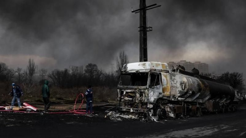 Trabajadores junto a un camión tanque destruido en un ataque a un depósito de petróleo. El cielo está negro con un humo espeso.