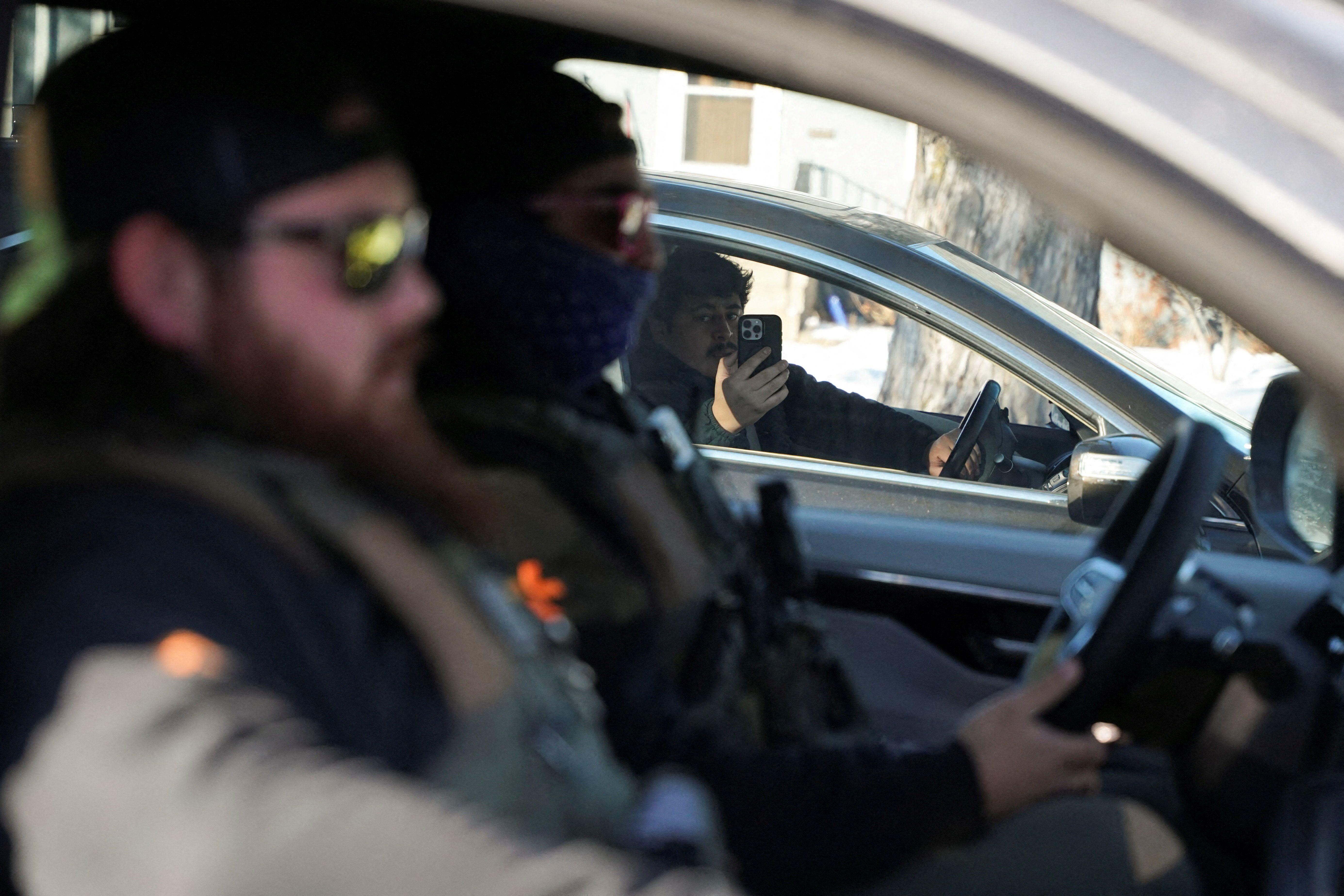 Un ciudadano al volante de un auto enfoca con su teléfono a dos agentes a bordo de otro auto en paralelo al suyo. Uno de ellos va encapuchado. 
