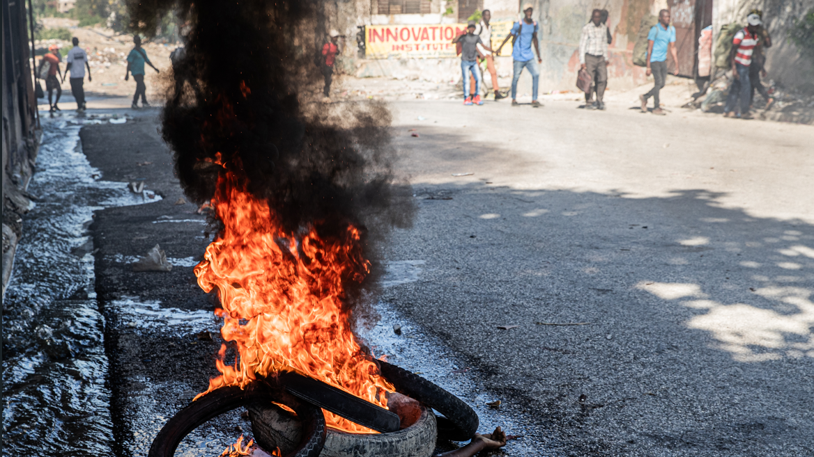 Cad&aacute;ver arde sob pneus em uma rua do centro de Porto Pr&iacute;ncipe, no dia 9 de fevereiro de 2026, com pessoas desfocadas ao fundo