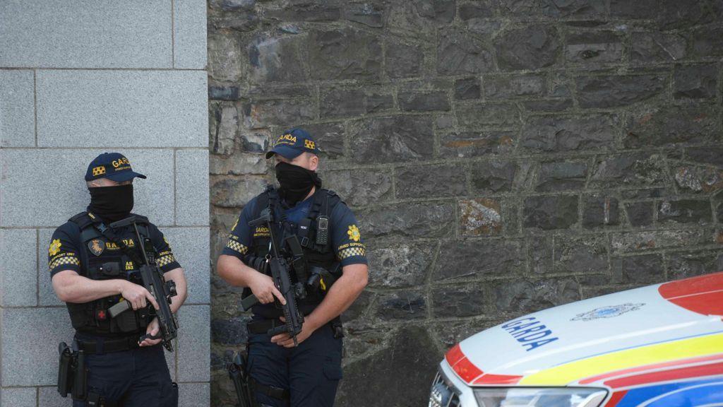 Dois guardas armados, usando uniforme e m&aacute;scaras, ao lado de um carro de pol&iacute;cia