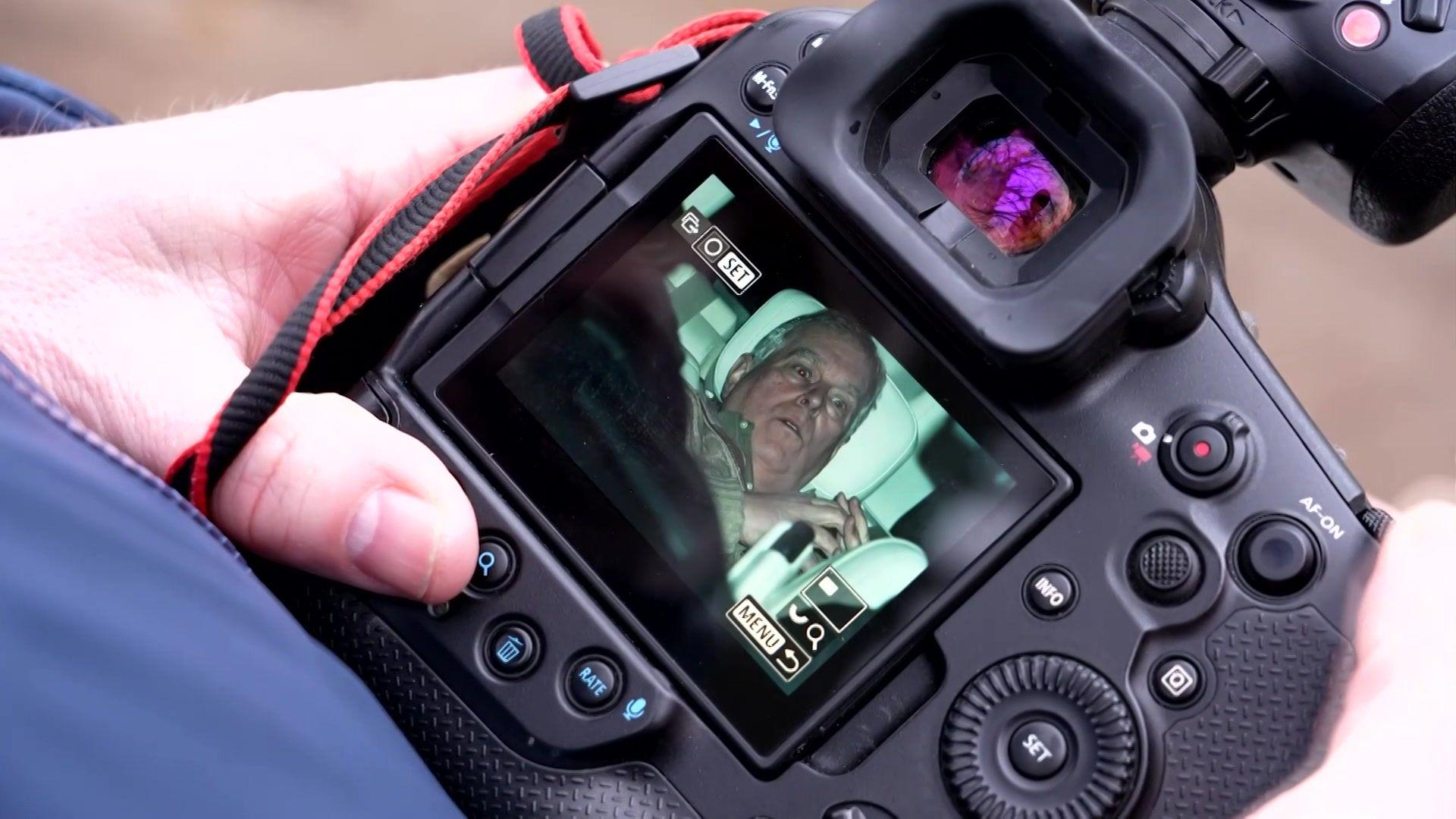 A man's hand holds a camera. On the display, an image of a shocked Andrew leaning back on a car seat can be seen